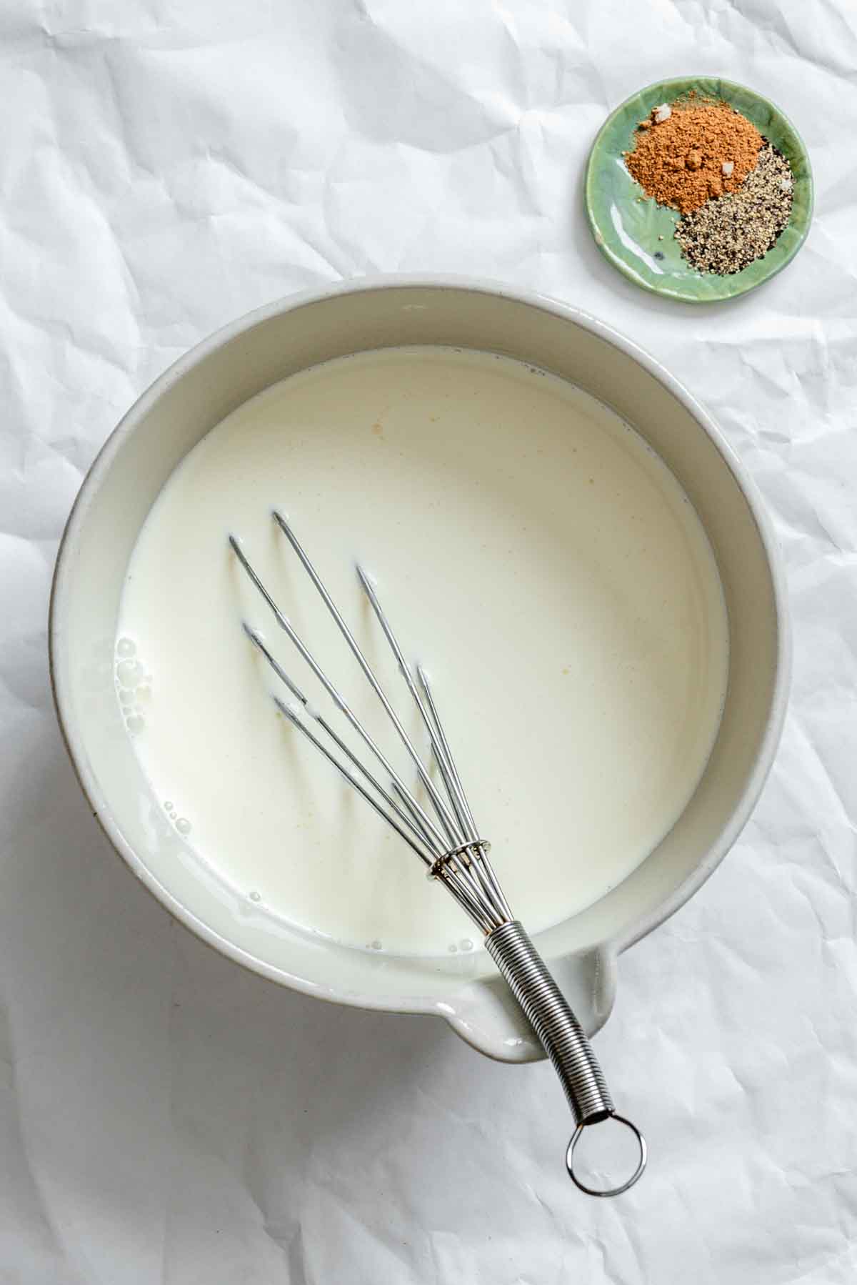 A bowl of creamy white béchamel sauce with a metal whisk, alongside a small green plate containing nutmeg and black pepper, all arranged on white parchment paper - the foundation ingredients for a gratin.