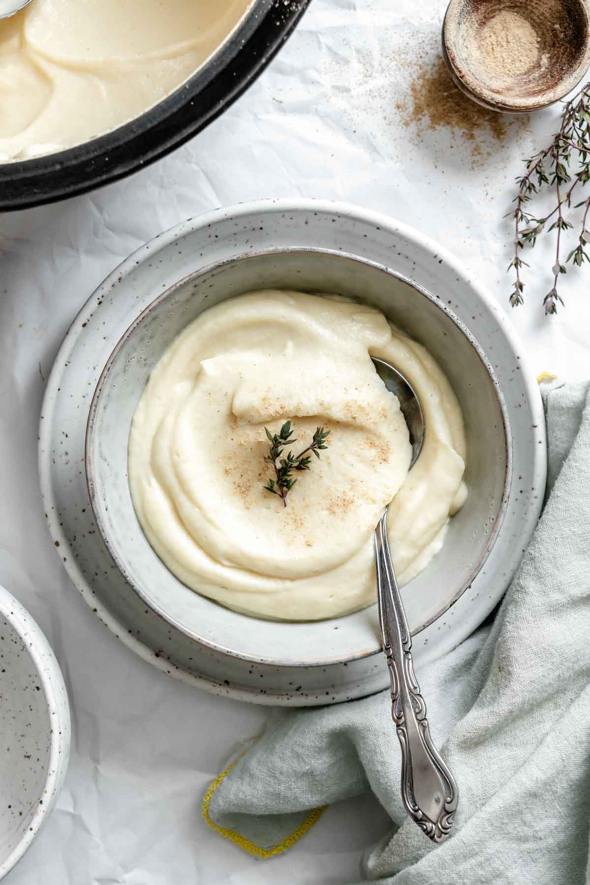 Celery root and parsnip puree served in a speckled ceramic bowl, garnished with fresh thyme and a light dusting of spice. A silver spoon rests in the creamy mixture.
