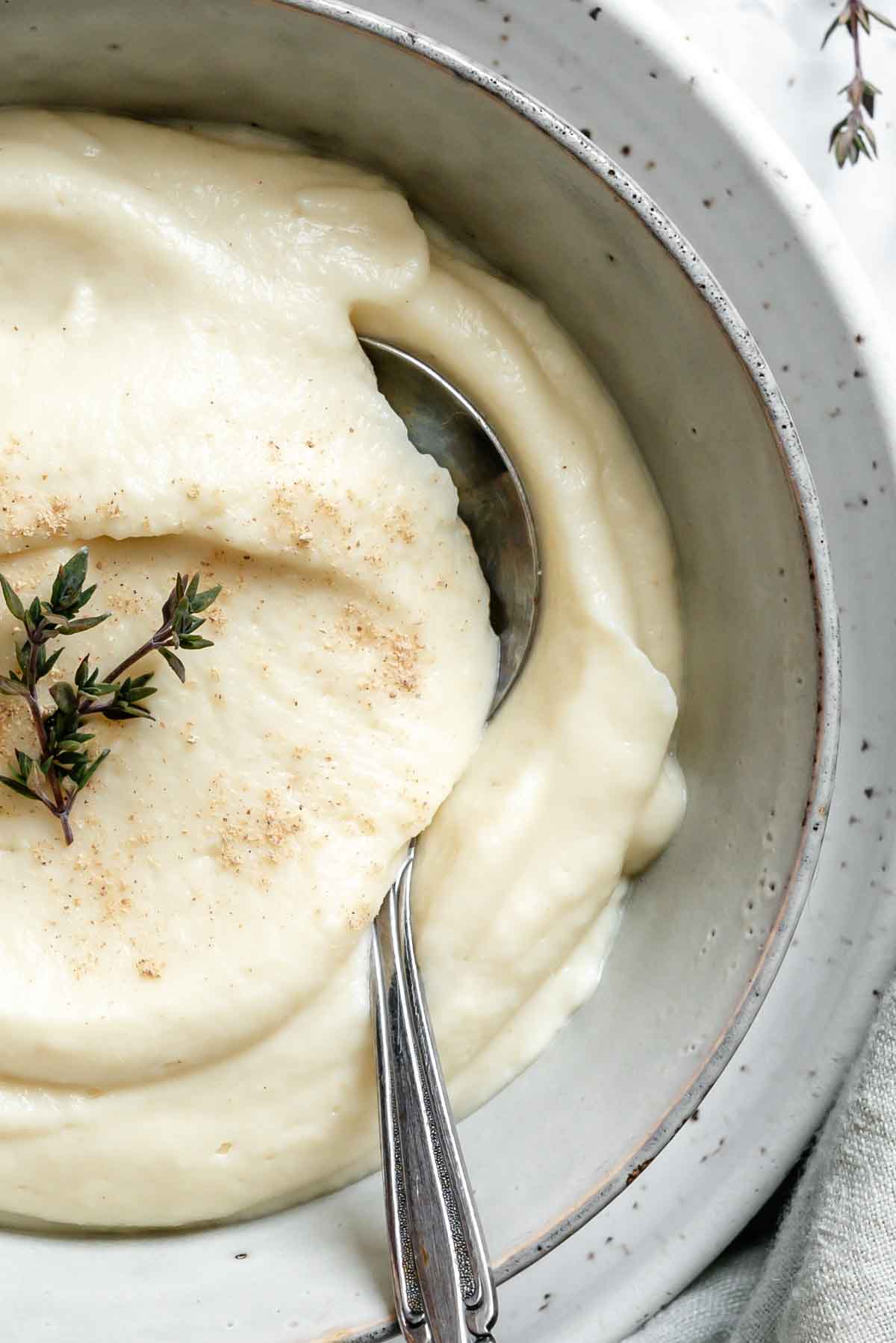 Close-up of celery root and parsnip puree in a white ceramic bowl. The silky smooth texture shows swirls from a spoon, garnished with fresh thyme and spices.