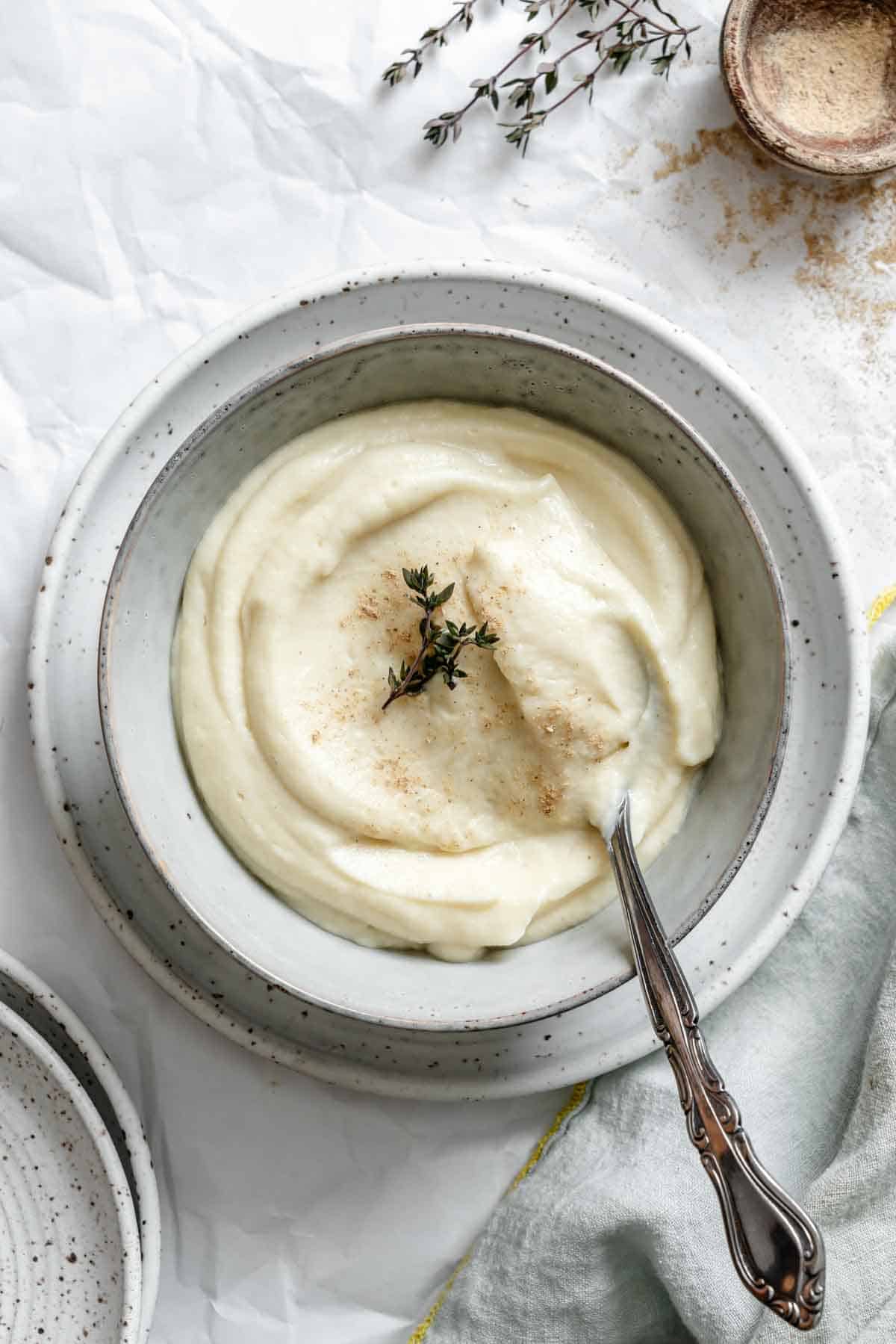 Overhead view of celery root and parsnip puree in a speckled ceramic bowl with vintage silverware. The smooth, pale puree is garnished with fresh herbs and spices on a white background.