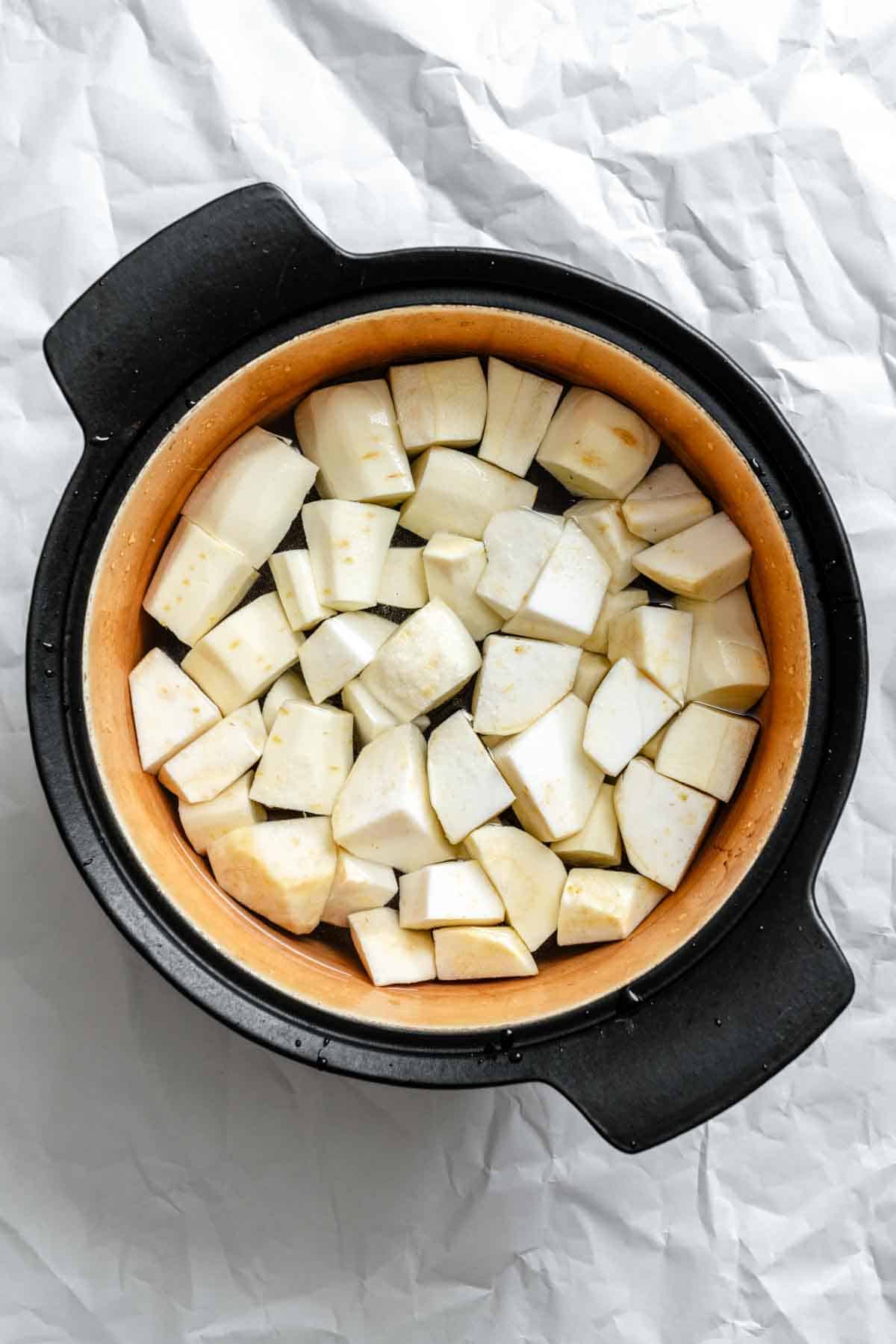 Raw parsnip and celery root pieces arranged in a black ceramic Dutch oven or pot on white background, ready to be cooked.