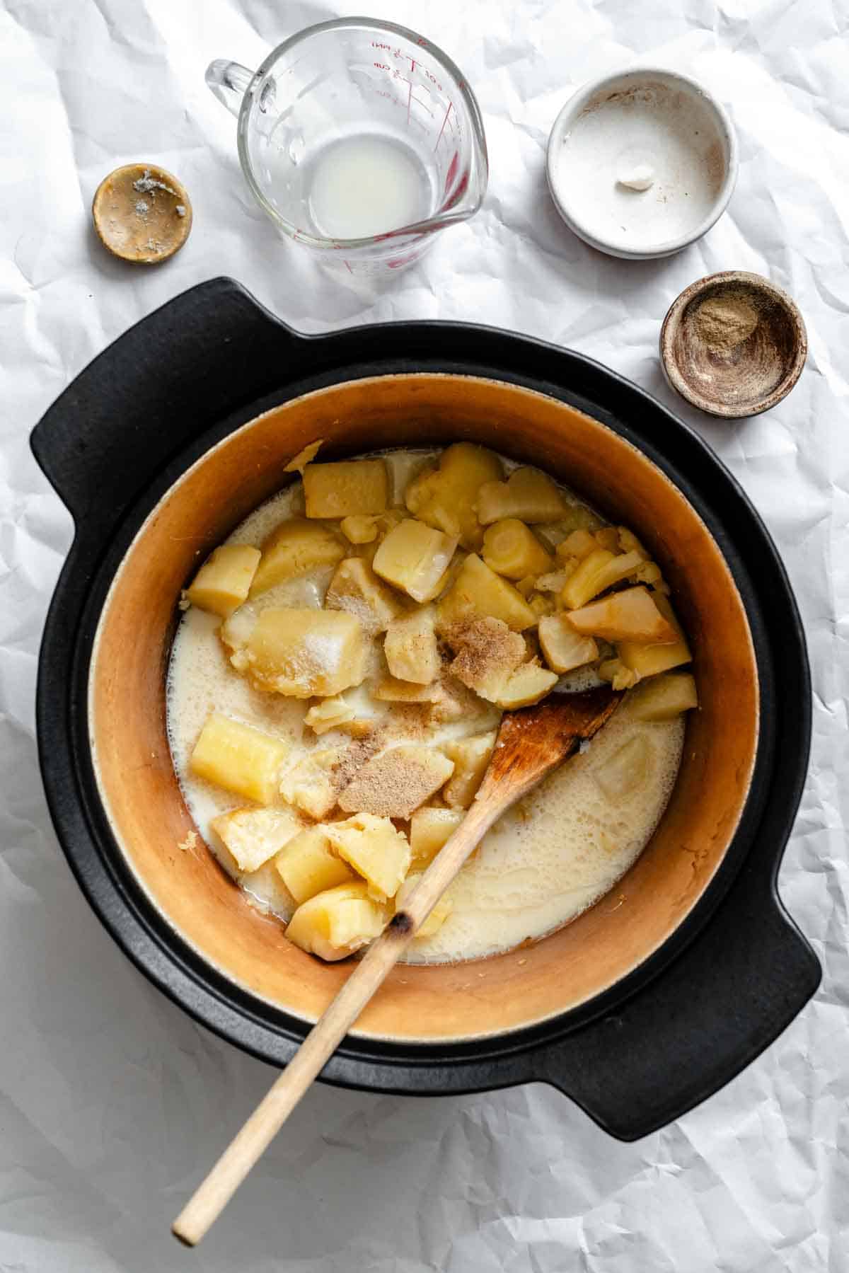 Cooking process of celery root and parsnip puree in a black Dutch oven. The root vegetables are simmering in liquid with spices and herbs, with a wooden spoon stirring the mixture.
