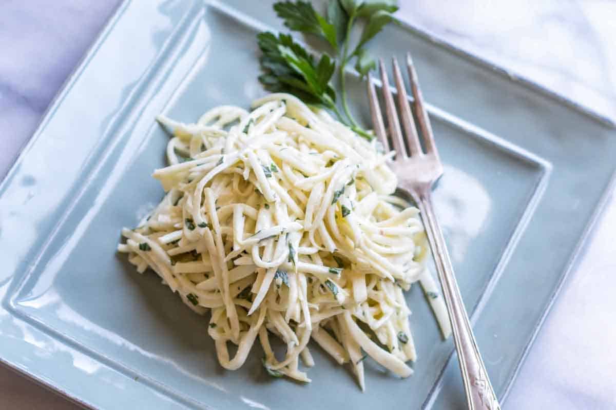 A different angle of the same celery root remoulade dish on the light blue square plate, set against a white marble surface. The julienned vegetable strands are dressed in a creamy sauce and mixed with fresh herbs, with a silver fork partially inserted in the dish.