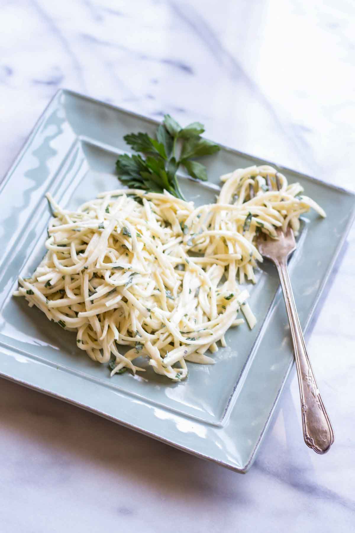 A light blue square plate containing julienned celery root remoulade with finely chopped parsley mixed throughout. The pale, creamy strands are garnished with a fresh parsley sprig, and a silver fork rests alongside the dish.