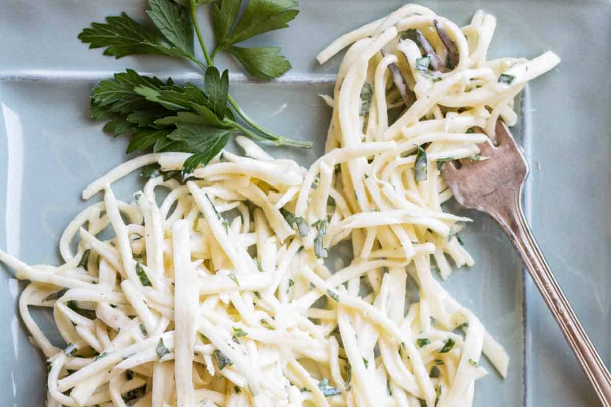A close-up view of the celery root remoulade on a light blue plate, showing the thin, matchstick-cut vegetable pieces mixed with herbs. Fresh parsley sprigs are arranged beside the salad, and a silver fork is visible on the right side.