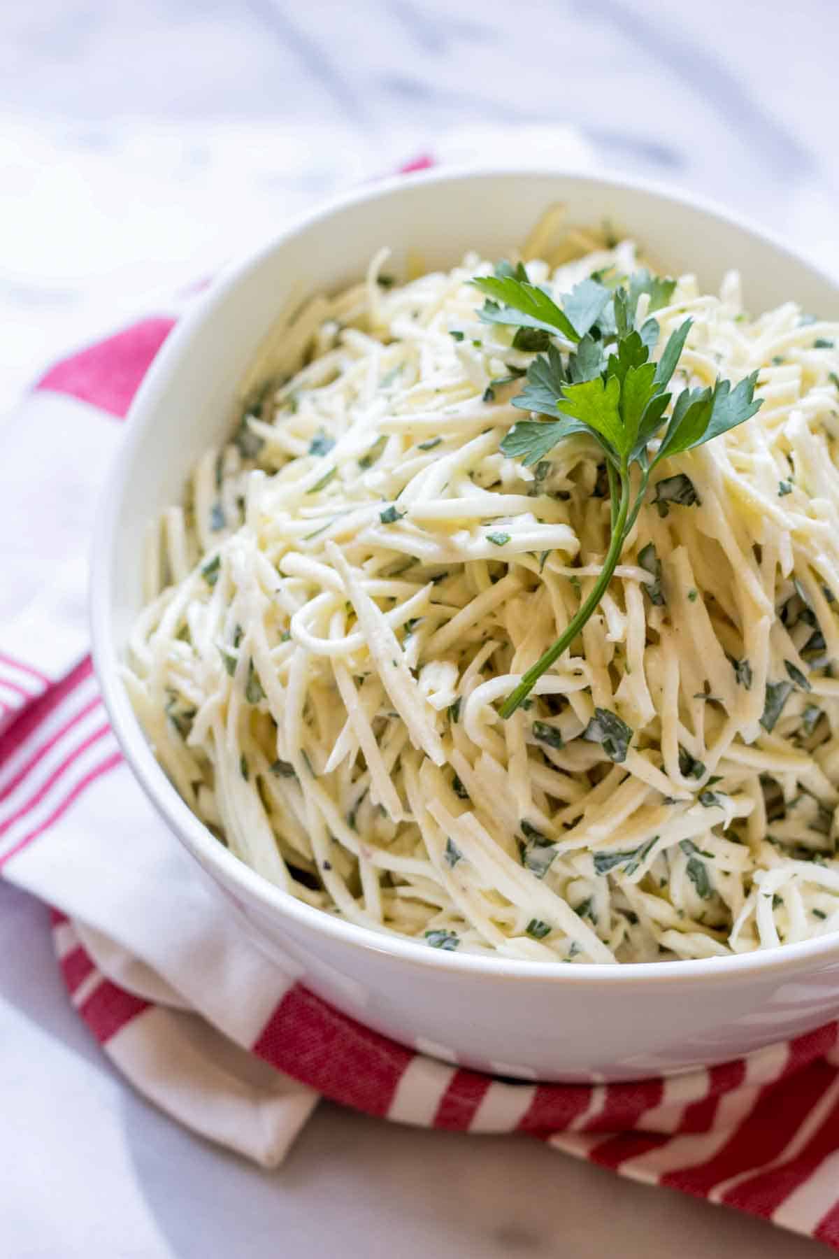 A white bowl filled with celery root remoulade garnished with a sprig of parsley on top. The creamy, herb-flecked salad is presented on a red and white striped cloth napkin against a white marble background.RetryClaude can make mistakes. Please double-check responses. 3.7 Sonnet
