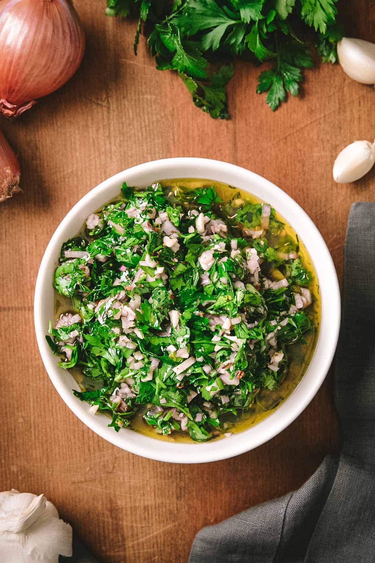 A white ceramic bowl of finished chimichurri sauce on a wooden board, showing the bright green herb mixture swimming in olive oil. Fresh parsley, shallots, and garlic cloves are visible in the background.