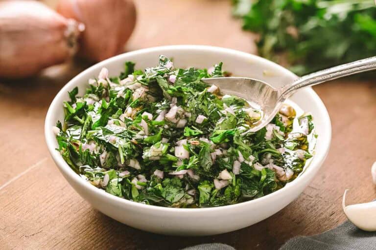 A close-up of a white bowl containing chimichurri sauce with a silver spoon, showcasing the texture of finely chopped herbs and red onions suspended in olive oil. In the background are steaks on a wooden board.