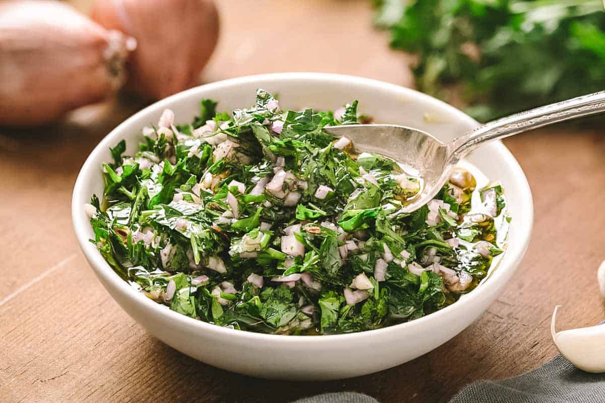 A close-up of a white bowl containing chimichurri sauce with a silver spoon, showcasing the texture of finely chopped herbs and red onions suspended in olive oil. In the background are steaks on a wooden board.