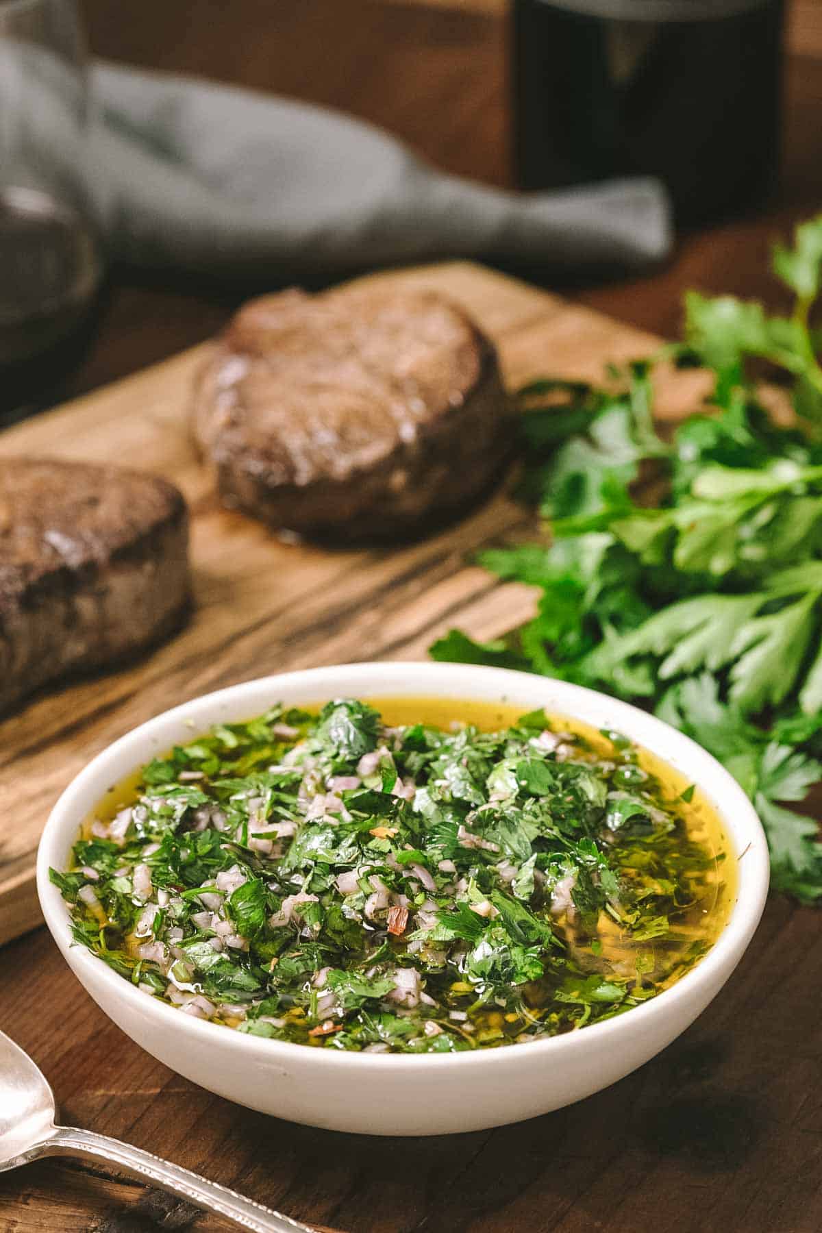 A white bowl of chimichurri sauce on a wooden surface with filet mignon steaks and fresh parsley in the background. The sauce appears shiny with olive oil and filled with finely chopped herbs.