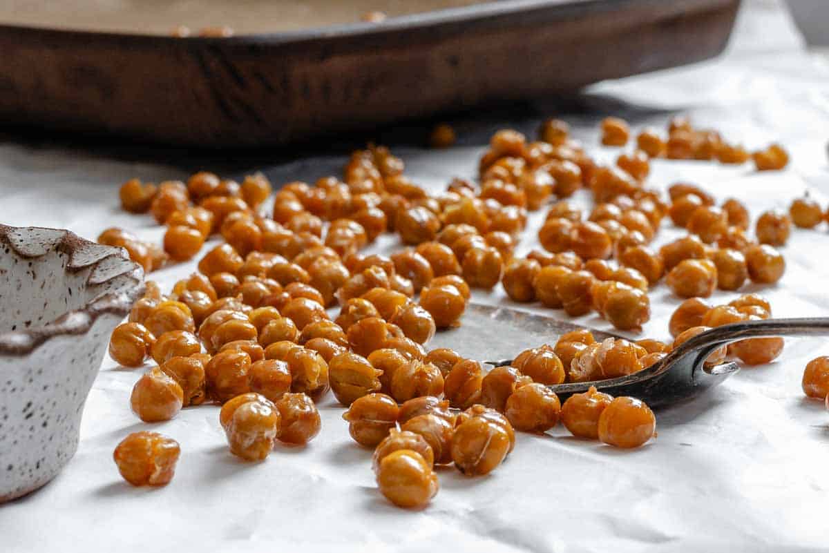 Roasted crispy chickpeas piled on white parchment paper with a silver fork and ceramic bowl visible in the background.