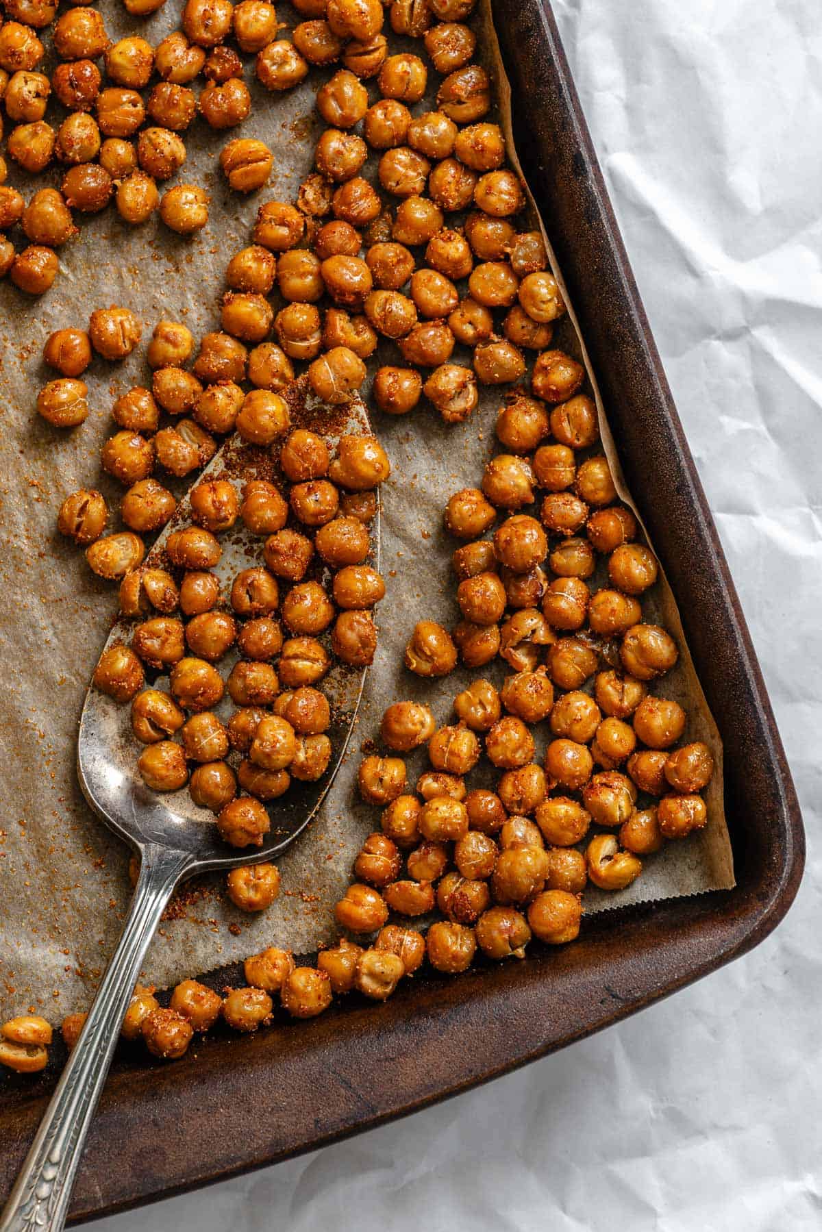 Close-up of roasted crispy chickpeas on a baking sheet with a serving spoon scooping some of the seasoned chickpeas.