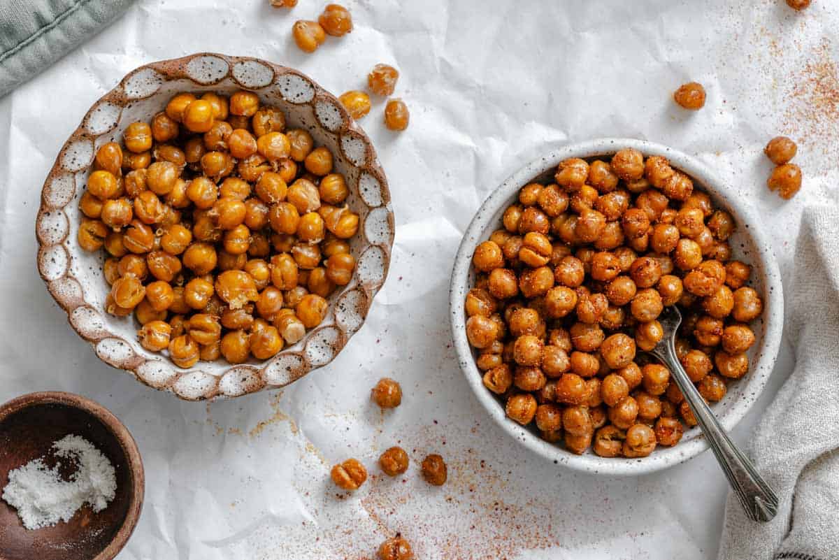 Overhead shot of two ceramic bowls filled with roasted seasoned chickpeas, with a small wooden bowl of salt nearby on white parchment paper.