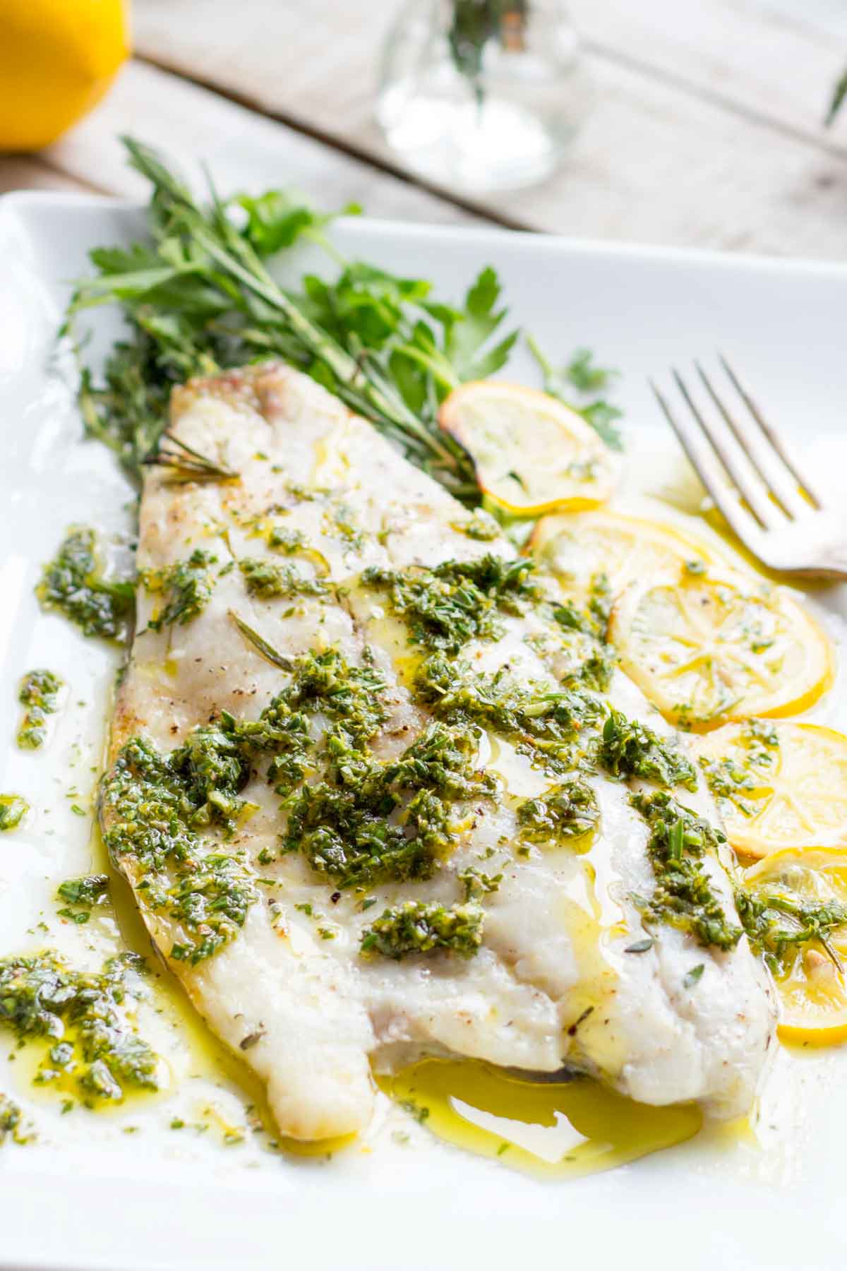 Close-up of cooked fish fillet on a white plate with lemon slices, herb sauce, and fresh parsley garnish.