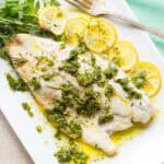 Overhead view of white fish fillet served on a rectangular plate with lemon slices, herb sauce, and fresh herbs.