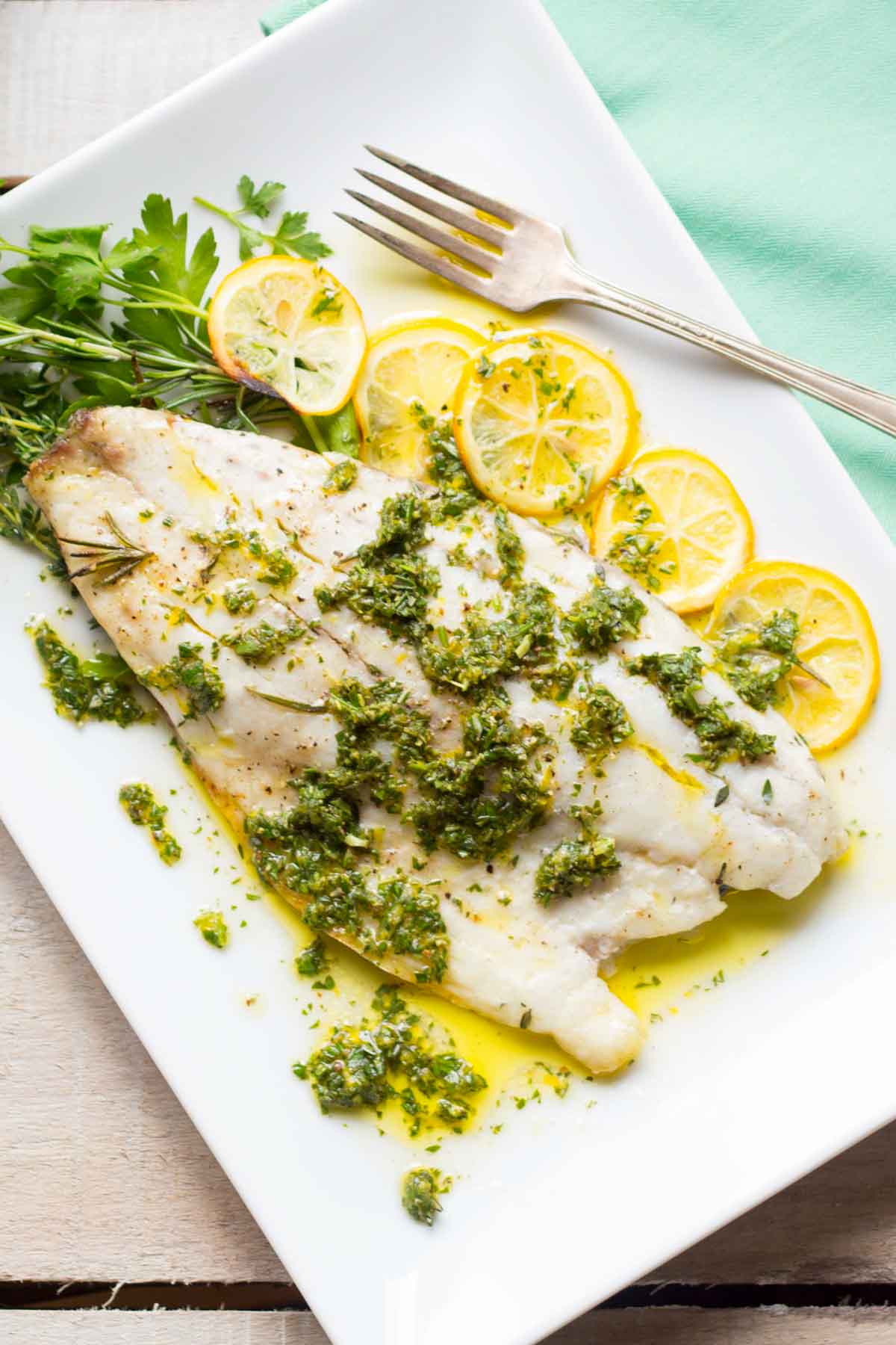 Overhead view of white fish fillet served on a rectangular plate with lemon slices, herb sauce, and fresh herbs.