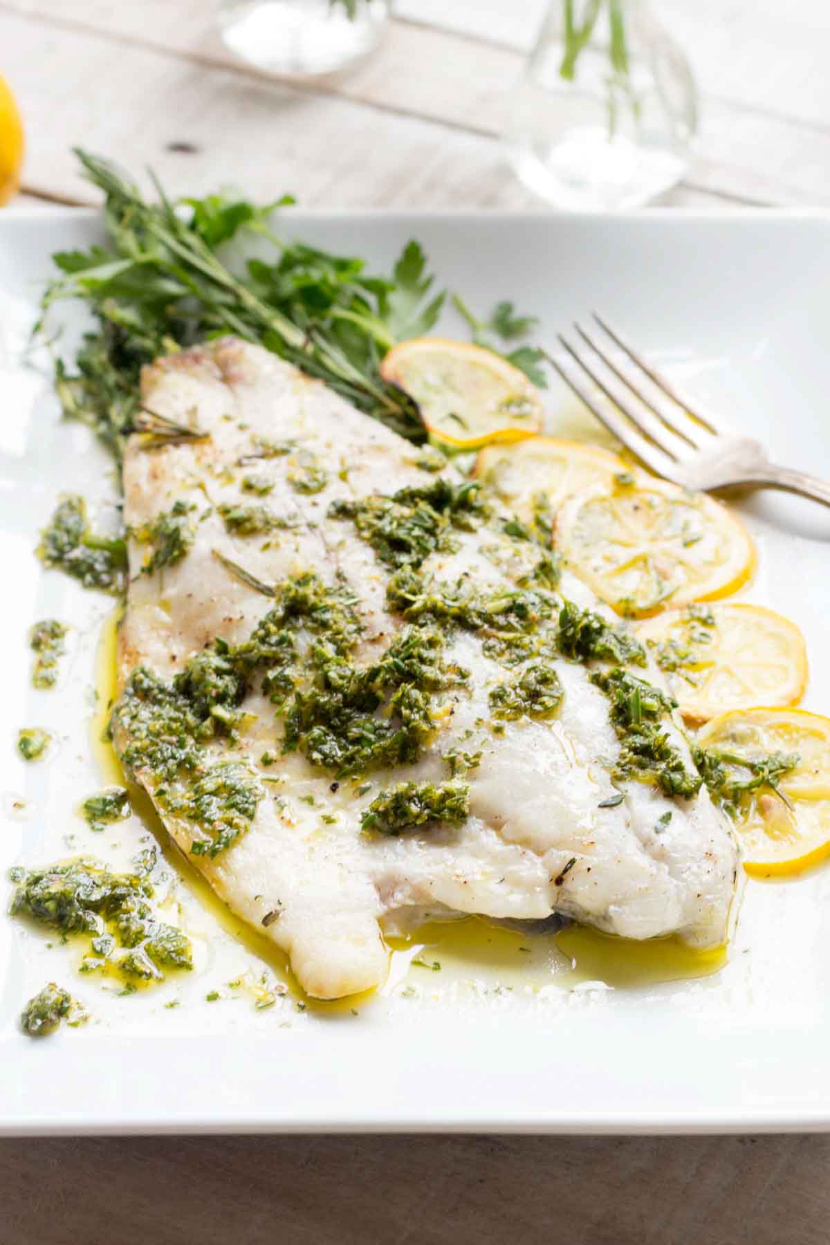 Similar angle of the plated fish with herb sauce, lemon slices and herbs on a white plate with turquoise napkin visible.