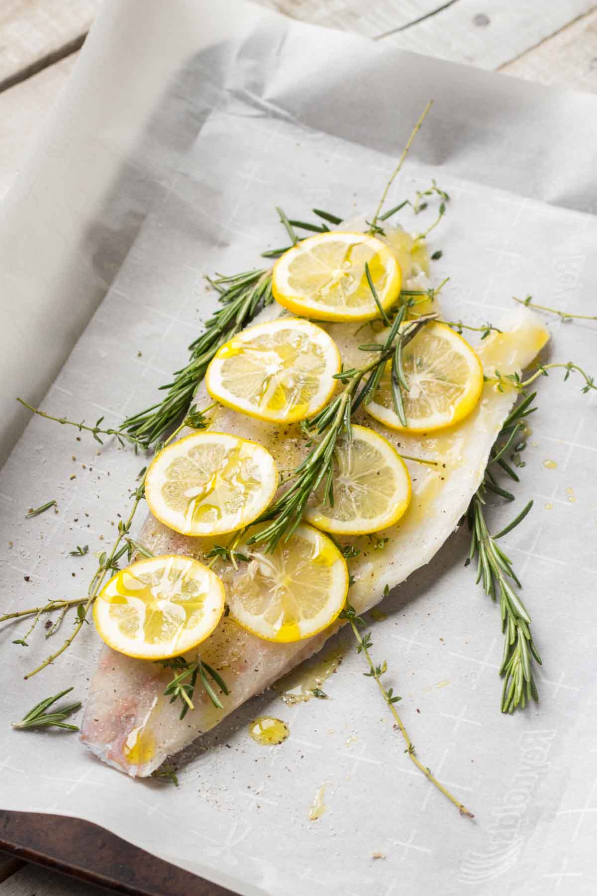 Raw fish fillet on parchment paper being prepared for baking, topped with lemon slices and fresh rosemary sprigs.