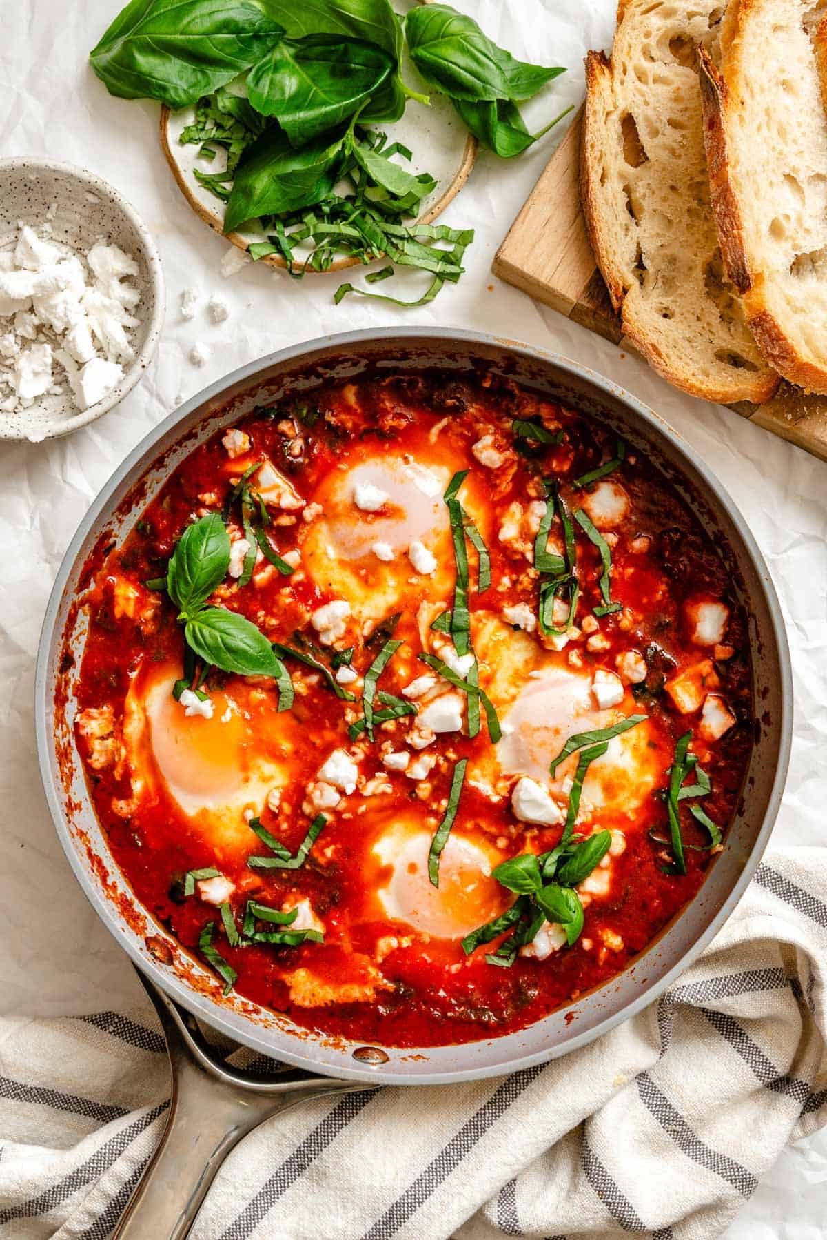 Overhead view of completed eggs in purgatory with poached eggs in spicy tomato sauce, garnished with fresh basil and crumbled goat cheese, served with crusty bread.