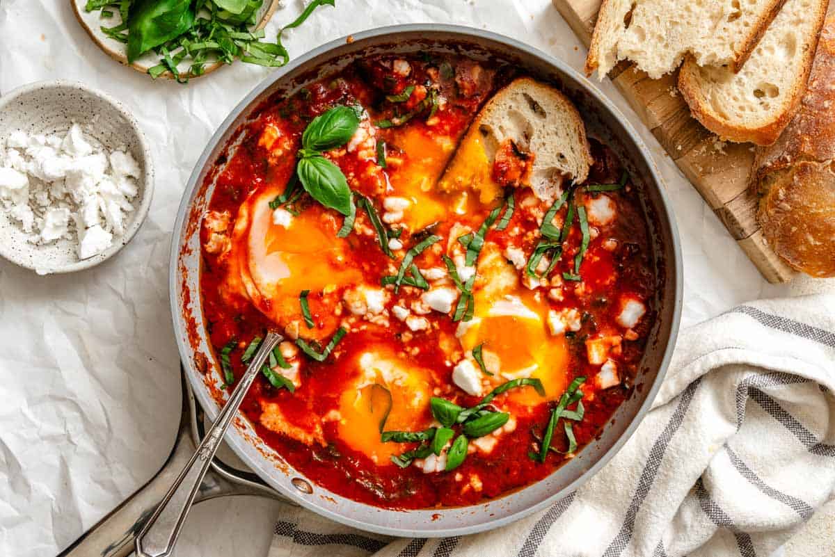 Finished eggs in purgatory in gray skillet with sliced rustic bread on wooden board, chopped basil and crumbled goat cheese in small bowls.