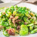 Plated fava bean dish garnished with chopped chives and prosciutto in a creamy sauce, served on a white plate with a spoon visible.