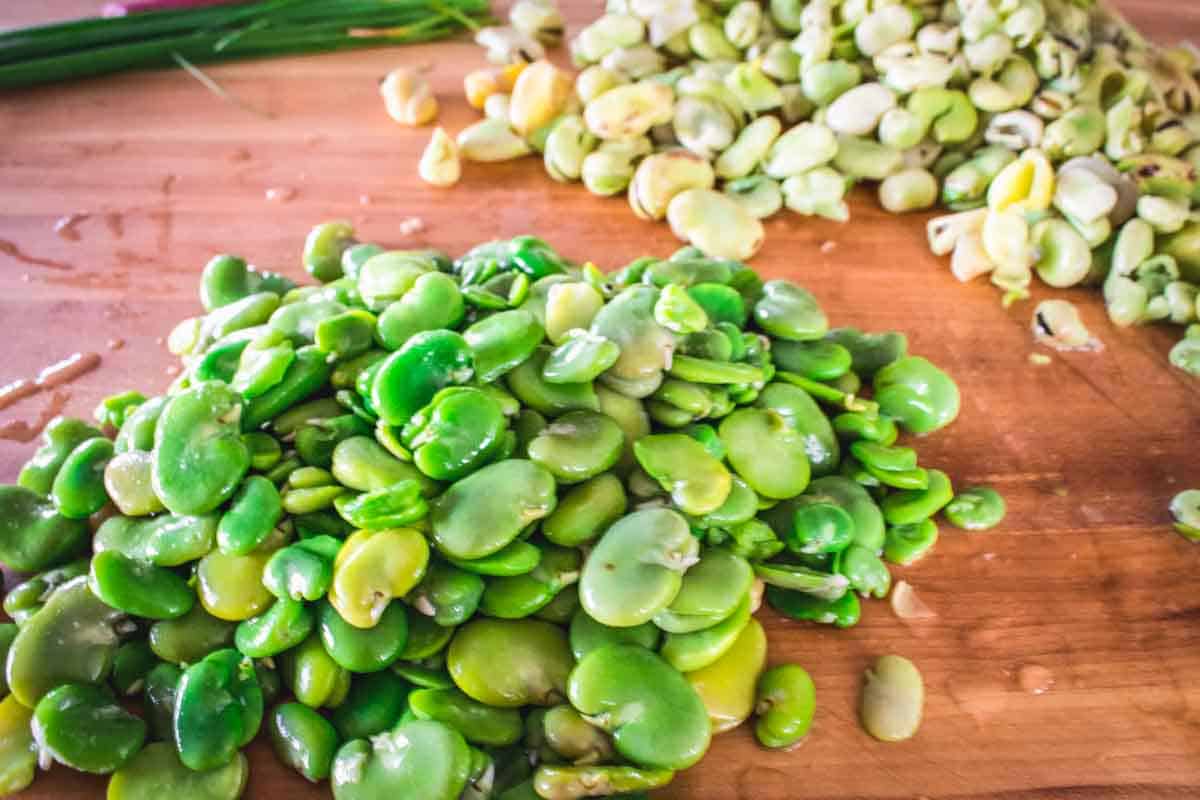 Fresh fava beans (broad beans) on a wooden cutting board, showing both shelled beans and some still in their pods, with green chives visible in the background.