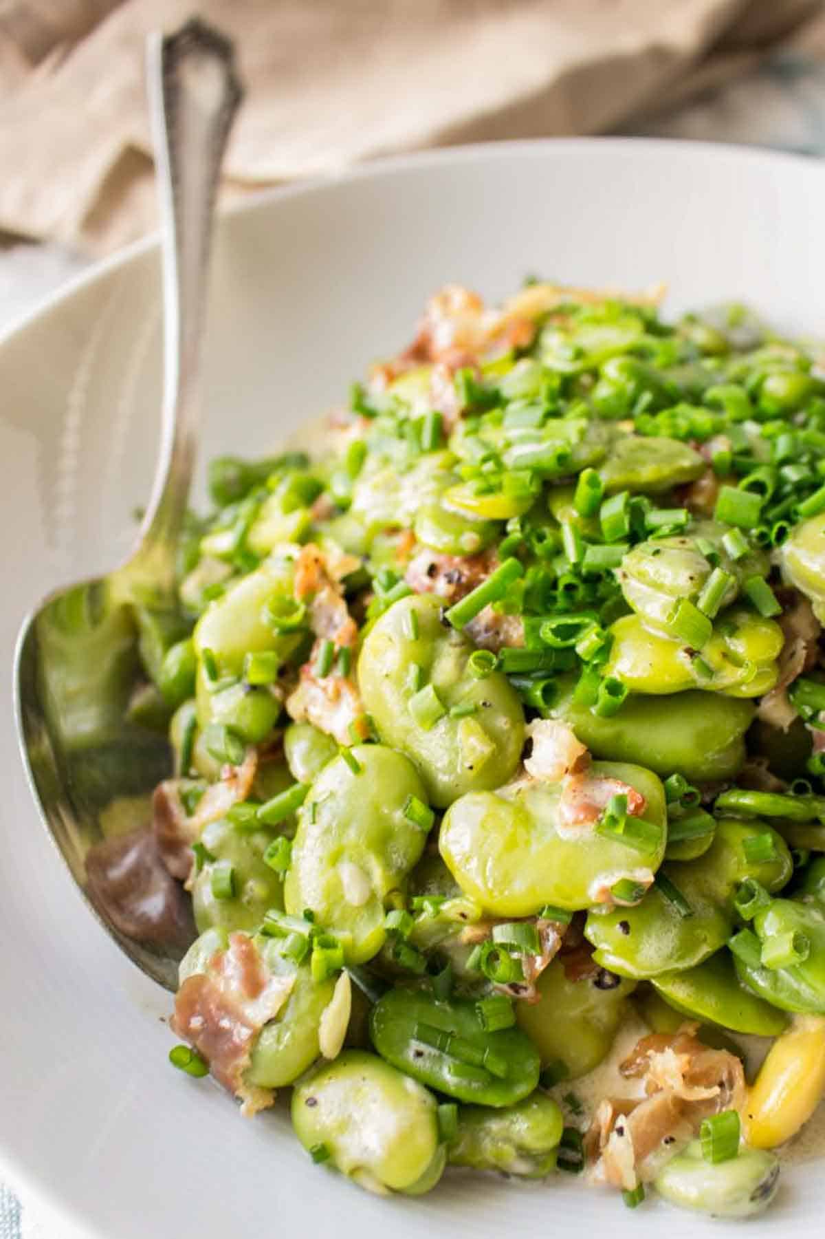 Close-up of a creamy fava bean dish with bits of prosciutto and chopped green onions in a white bowl with a serving spoon.
