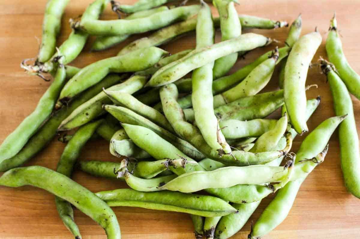 Whole fresh fava bean pods scattered on a wooden cutting board, showing their long green characteristic shape before being shelled.