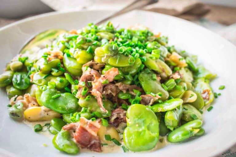 Plated fava bean dish garnished with chopped chives and prosciutto in a creamy sauce, served on a white plate with a spoon visible.