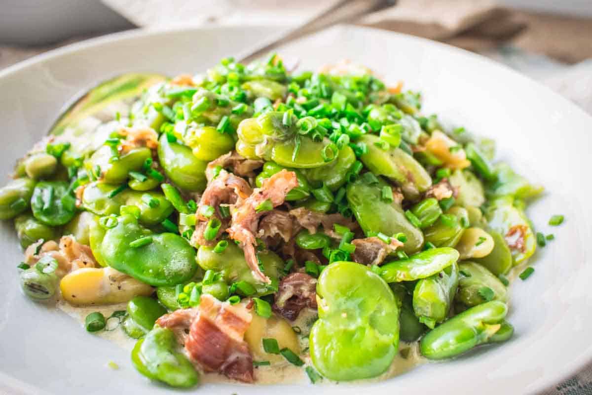 Plated fava bean dish garnished with chopped chives and prosciutto in a creamy sauce, served on a white plate with a spoon visible.