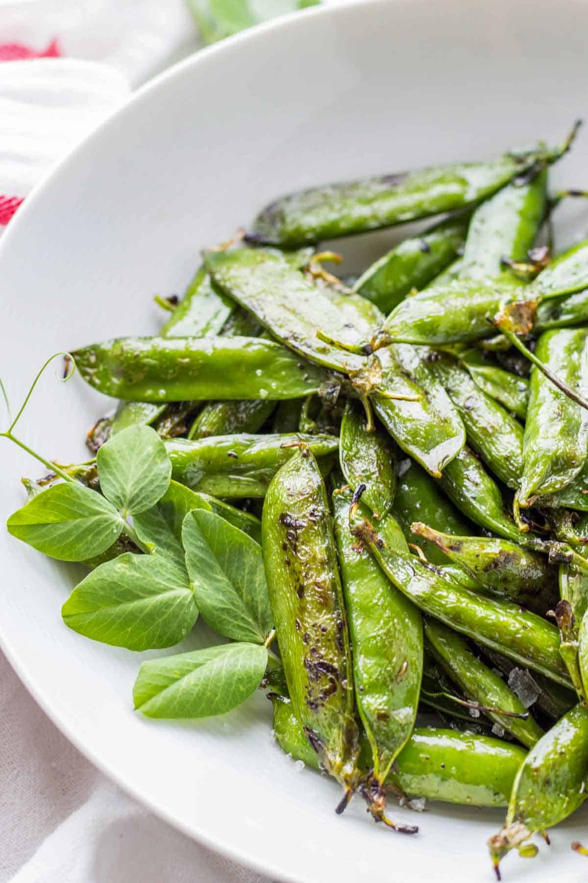 Top-down view of a white bowl filled with roasted sugar snap peas or English peas in their pods. The vibrant green pods have slight char marks and are garnished with fresh pea shoots and tendrils. A red-striped kitchen towel is visible in the background.