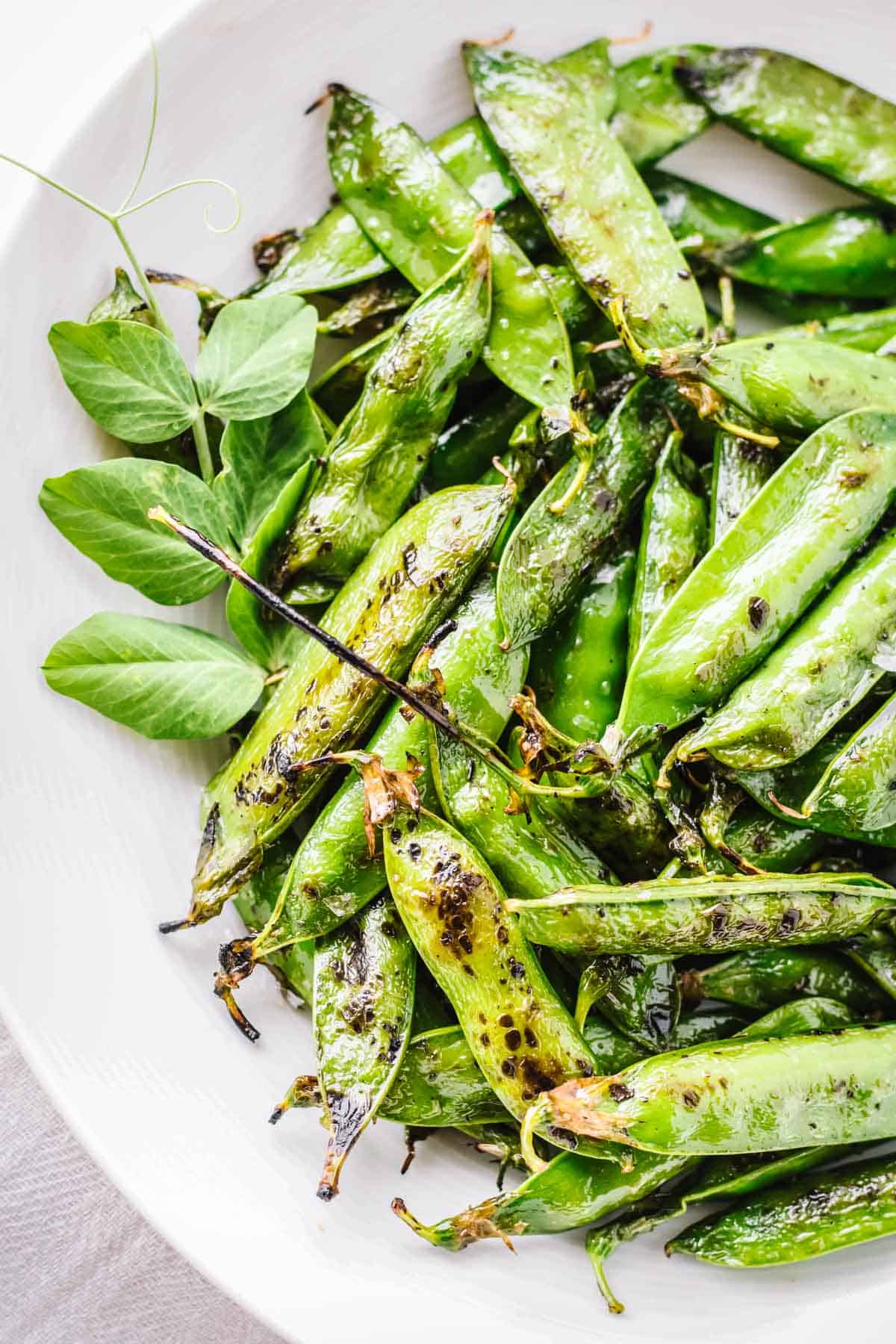 Overhead view of blistered green pea pods arranged on a white plate. The snap peas or English peas show caramelized edges from high-heat cooking with decorative pea tendrils and leaves as garnish. The bright green pods have a shiny, seasoned appearance.