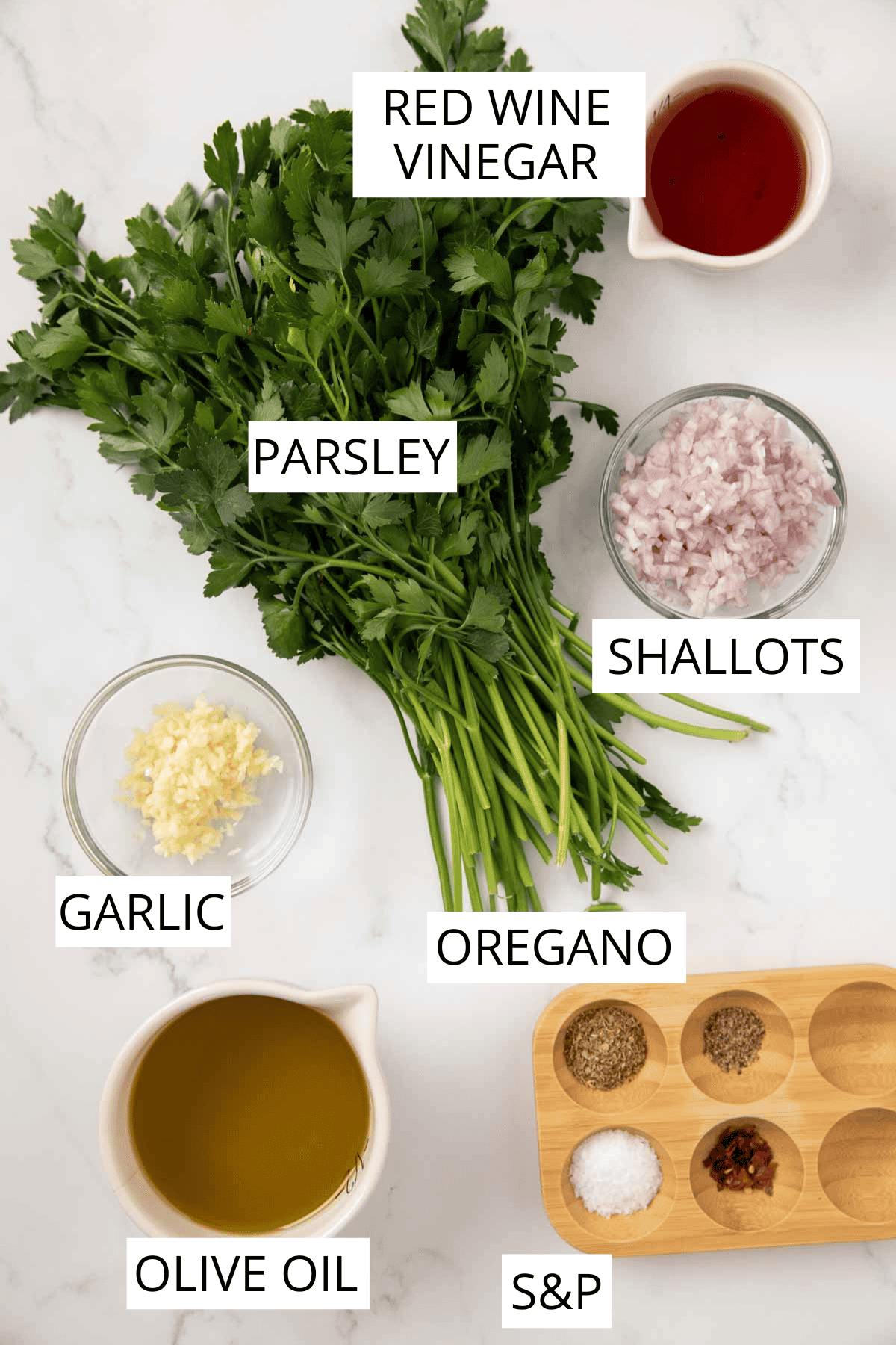 An overhead view of ingredients for chimichurri sauce arranged on white marble: a bunch of fresh parsley, bowls containing minced garlic and shallots, small pitchers of olive oil and red wine vinegar, and a wooden board with spices.