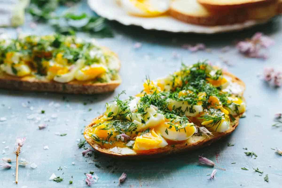 Several slices of toast topped with chopped soft-boiled eggs and fresh dill on a blue surface, with small purple flower petals scattered around.