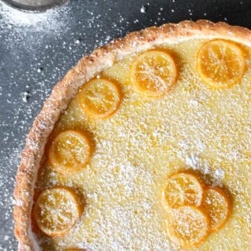 Closeup of a golden Meyer lemon tart with thin lemon slices arranged on a creamy yellow filling, dusted with powdered sugar. A small mesh sifter with powdered sugar is visible in the corner on a dark countertop.