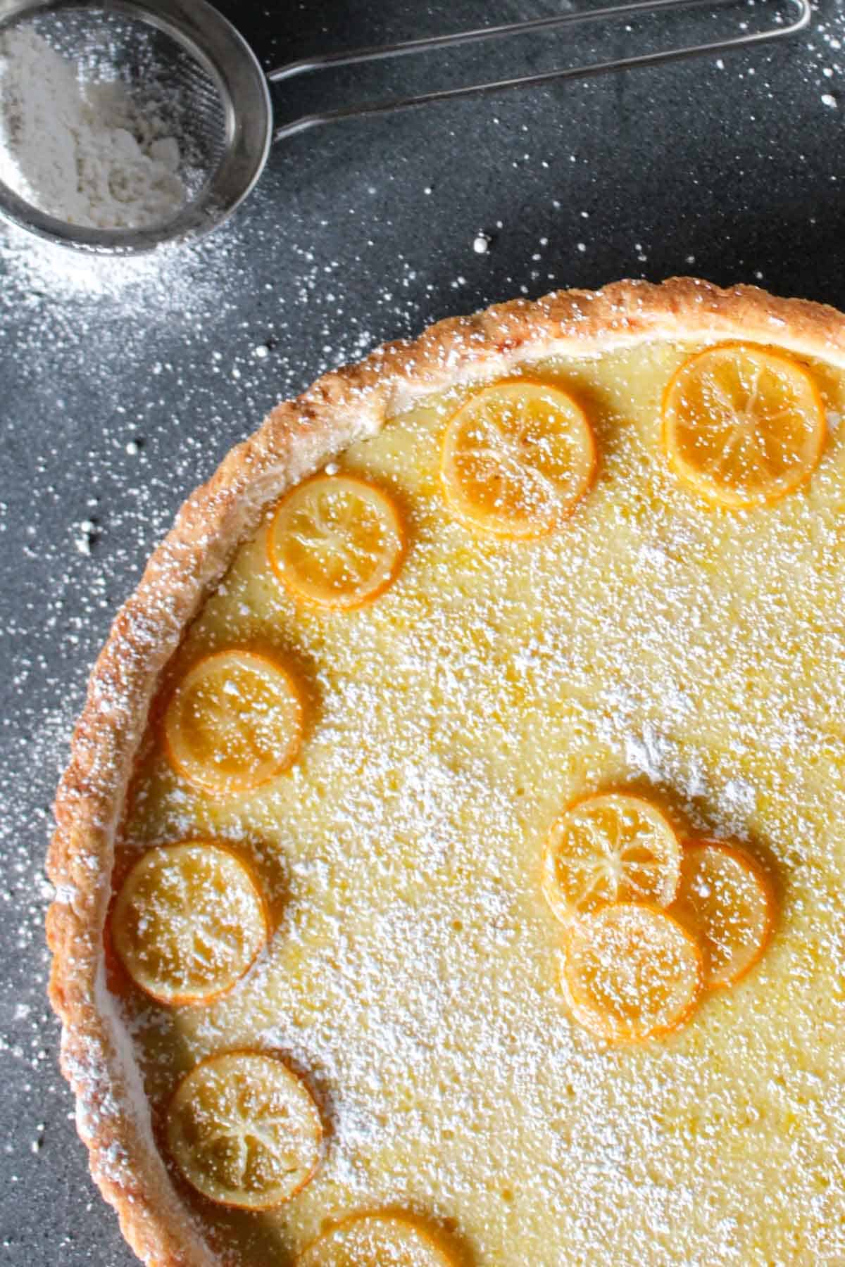 Closeup of a golden Meyer lemon tart with thin lemon slices arranged on a creamy yellow filling, dusted with powdered sugar. A small mesh sifter with powdered sugar is visible in the corner on a dark countertop.