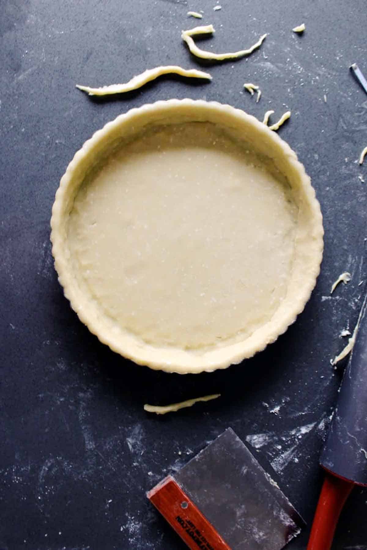 Overhead view of an unbaked tart shell in a fluted pan with pie dough trimmings scattered on a dark countertop. A pastry scraper with a red handle is visible at the bottom of the frame.