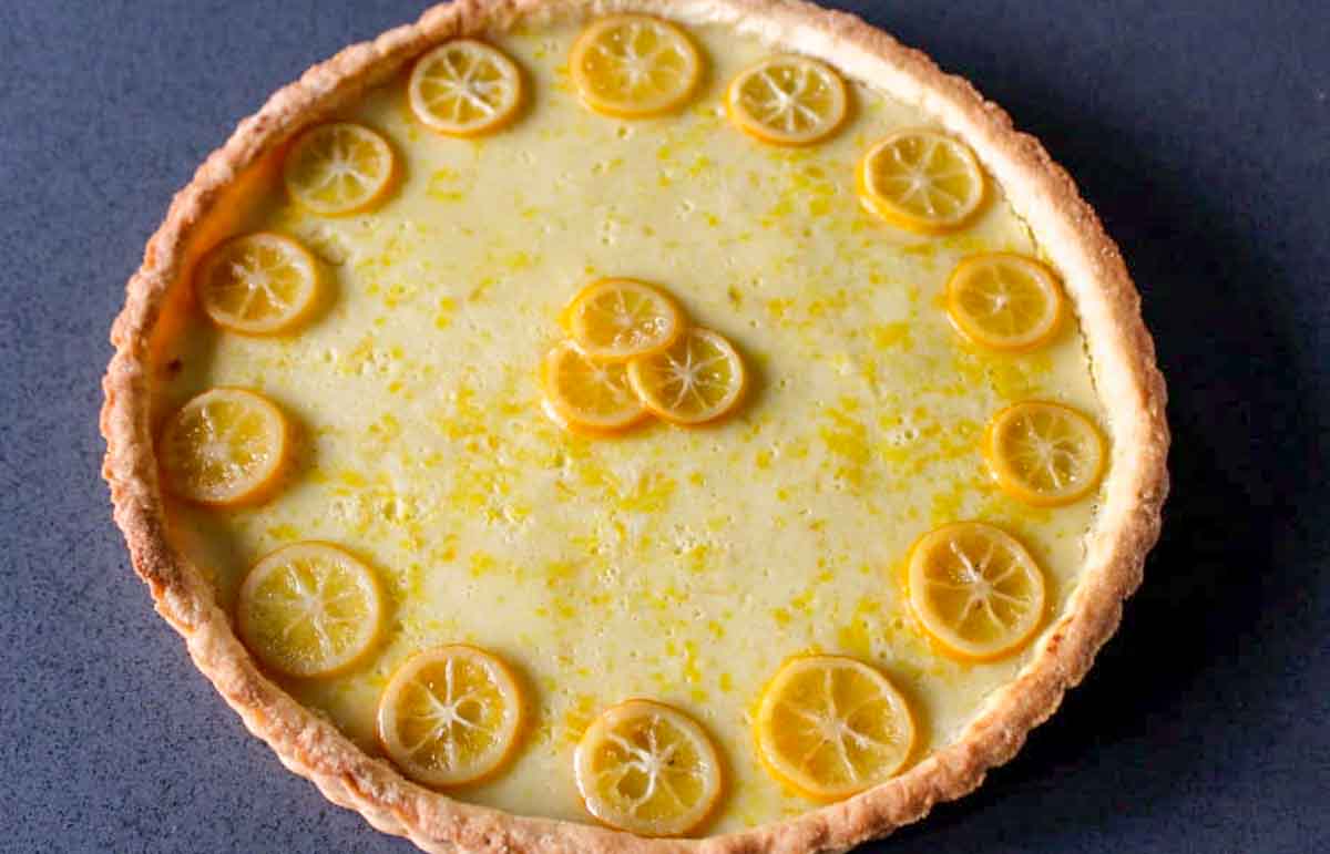 Top-down view of a baked Meyer lemon tart with thin lemon slices arranged in a circular pattern around the edge and clustered in the center on a yellow custard filling against a dark blue background.