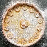 Overhead shot of a completed Meyer lemon tart generously dusted with powdered sugar, showing thin slices of lemon arranged decoratively around the edge and in the center, on a dark surface.