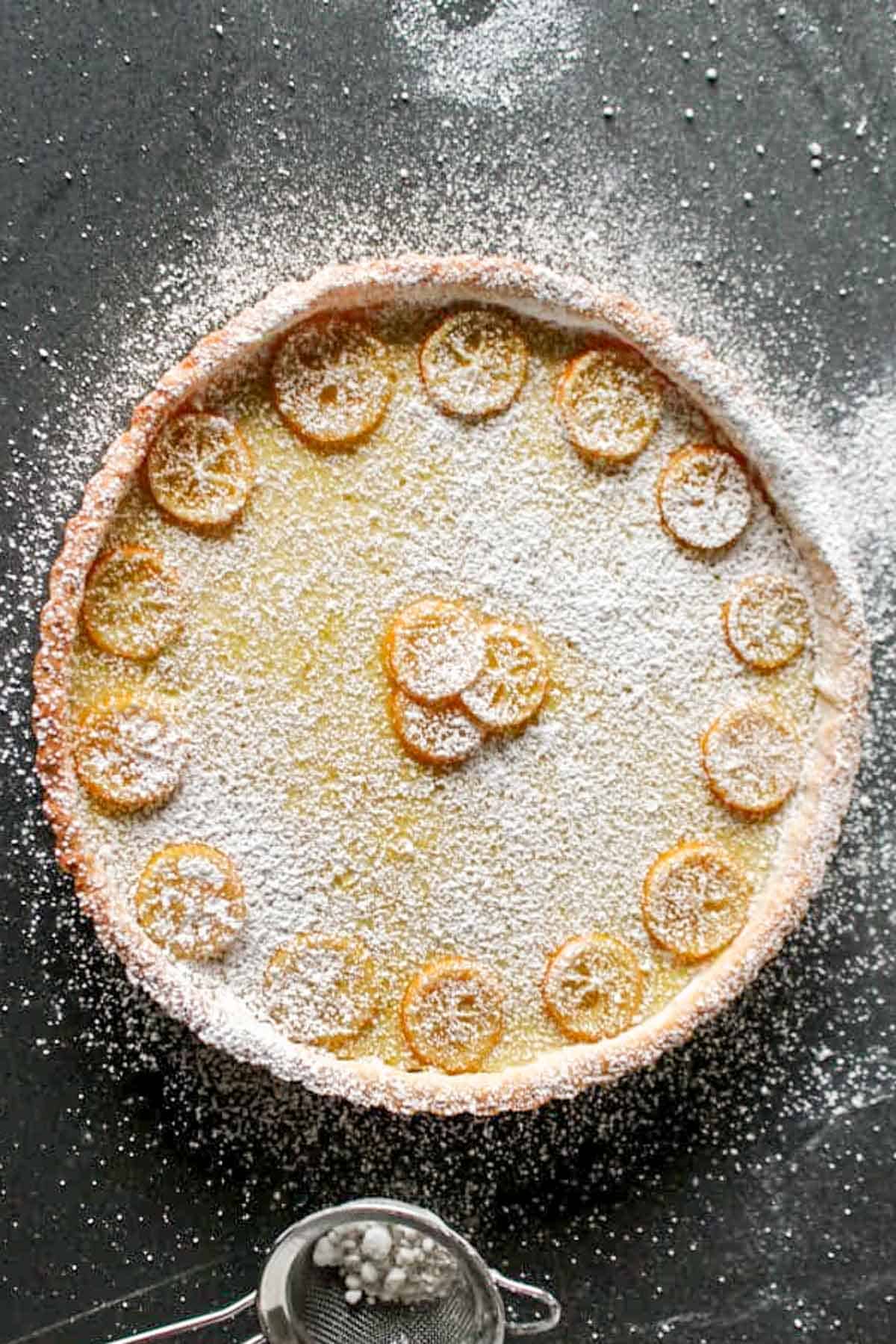 Overhead shot of a completed Meyer lemon tart generously dusted with powdered sugar, showing thin slices of lemon arranged decoratively around the edge and in the center, on a dark surface.
