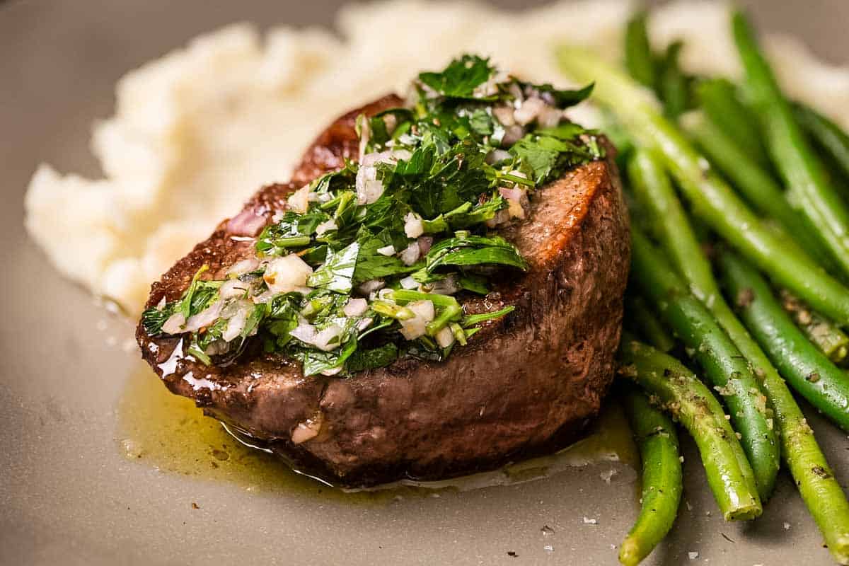 A filet mignon topped with a herb gremolata alongside green beans and mashed potatoes on a gray plate. The steak sits in a small pool of sauce or melted butter.