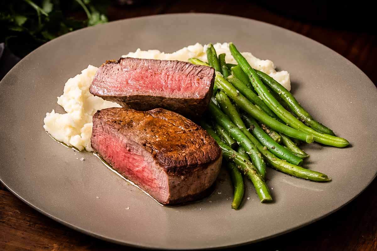 Two medium-rare filet mignon medallions served with mashed potatoes and green beans on a matte gray plate. The steaks are cut to show their pinkish-red centers against the browned exterior.