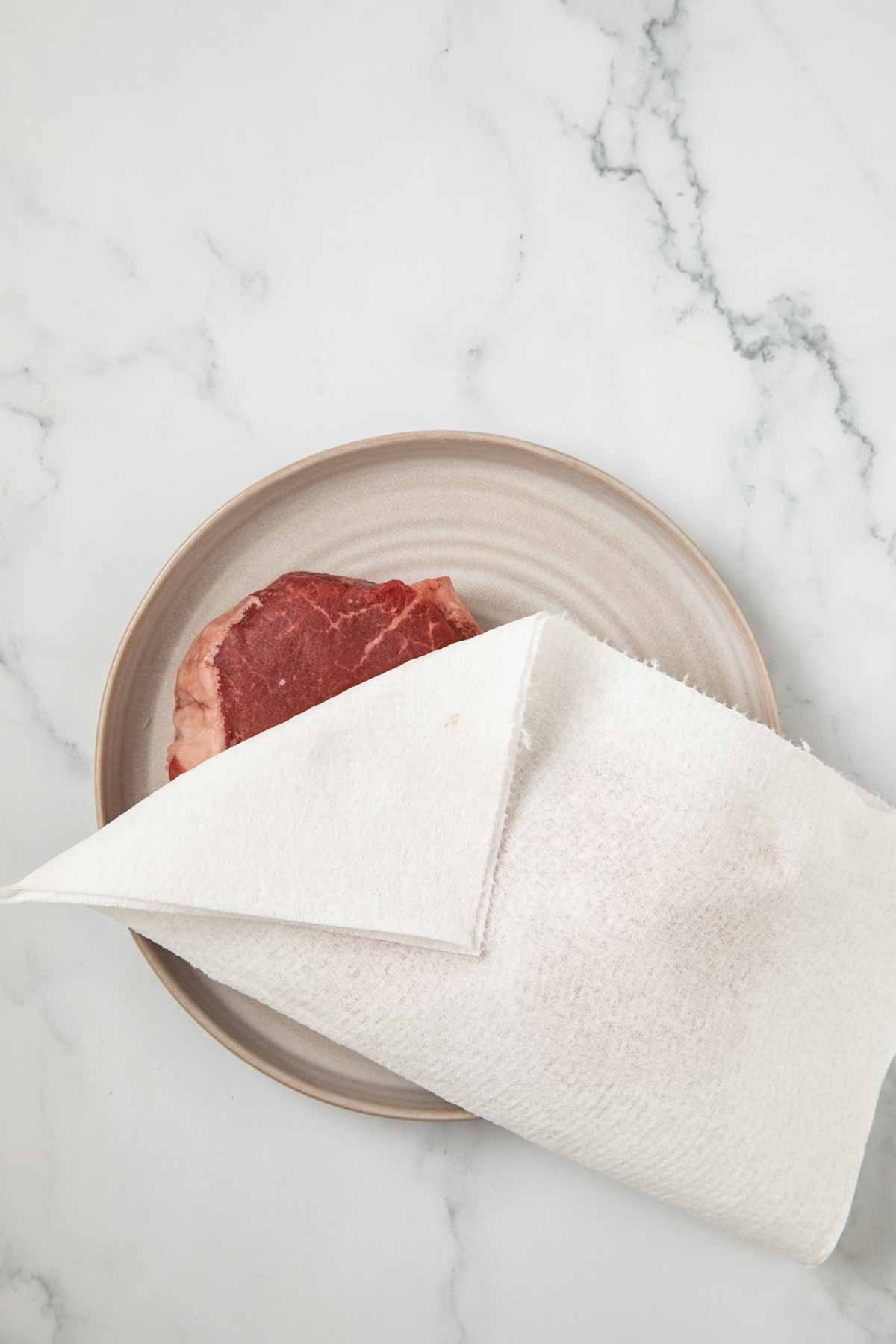 A light beige ceramic plate on a white marble countertop containing a raw beef filet partially covered with a white paper towel. The red meat is visible with marbling characteristic of a high-quality cut.