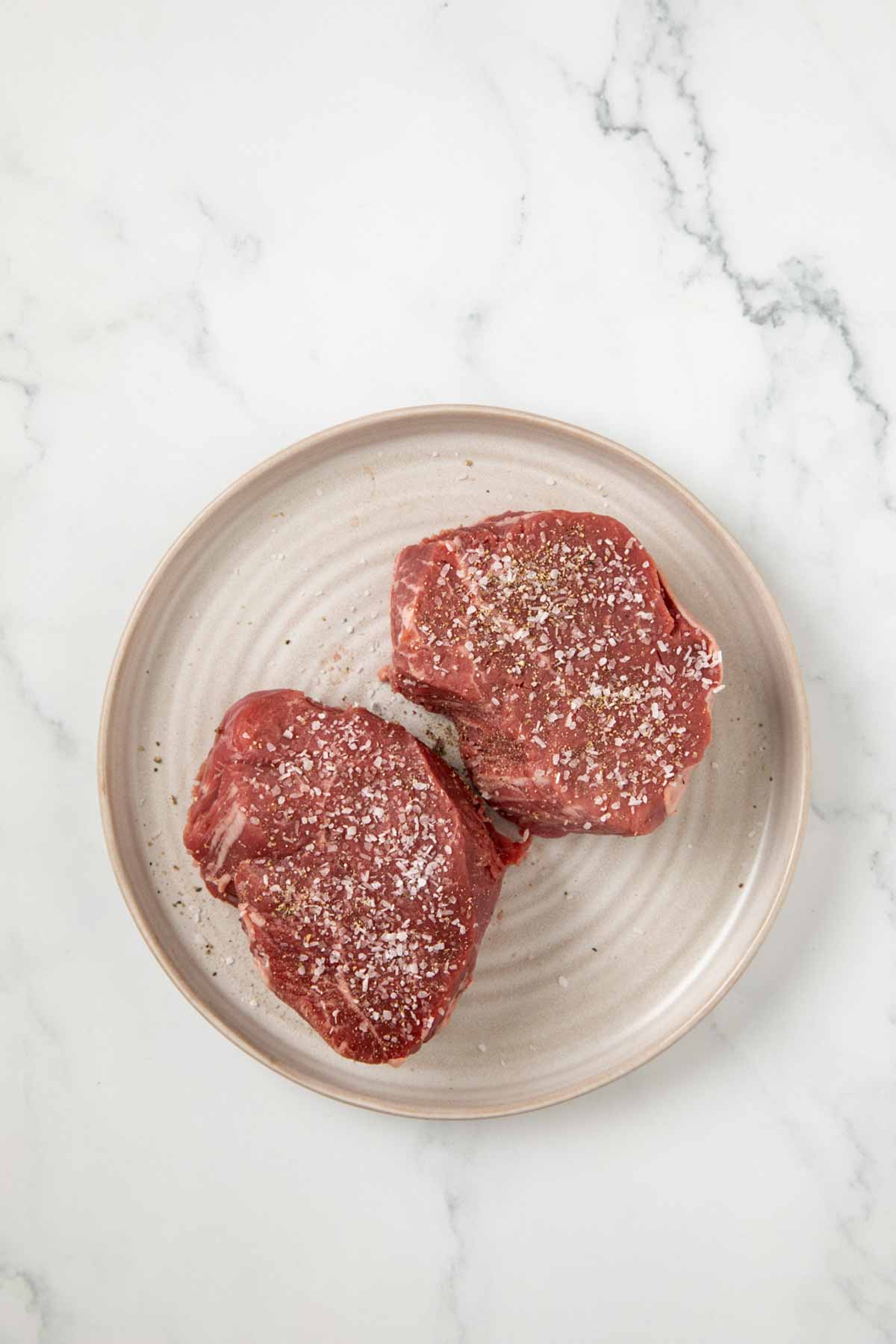 Two raw filet mignon steaks seasoned with salt and pepper on a beige ceramic plate against a white marble background. The steaks display the characteristic round, thick shape of filet cuts.