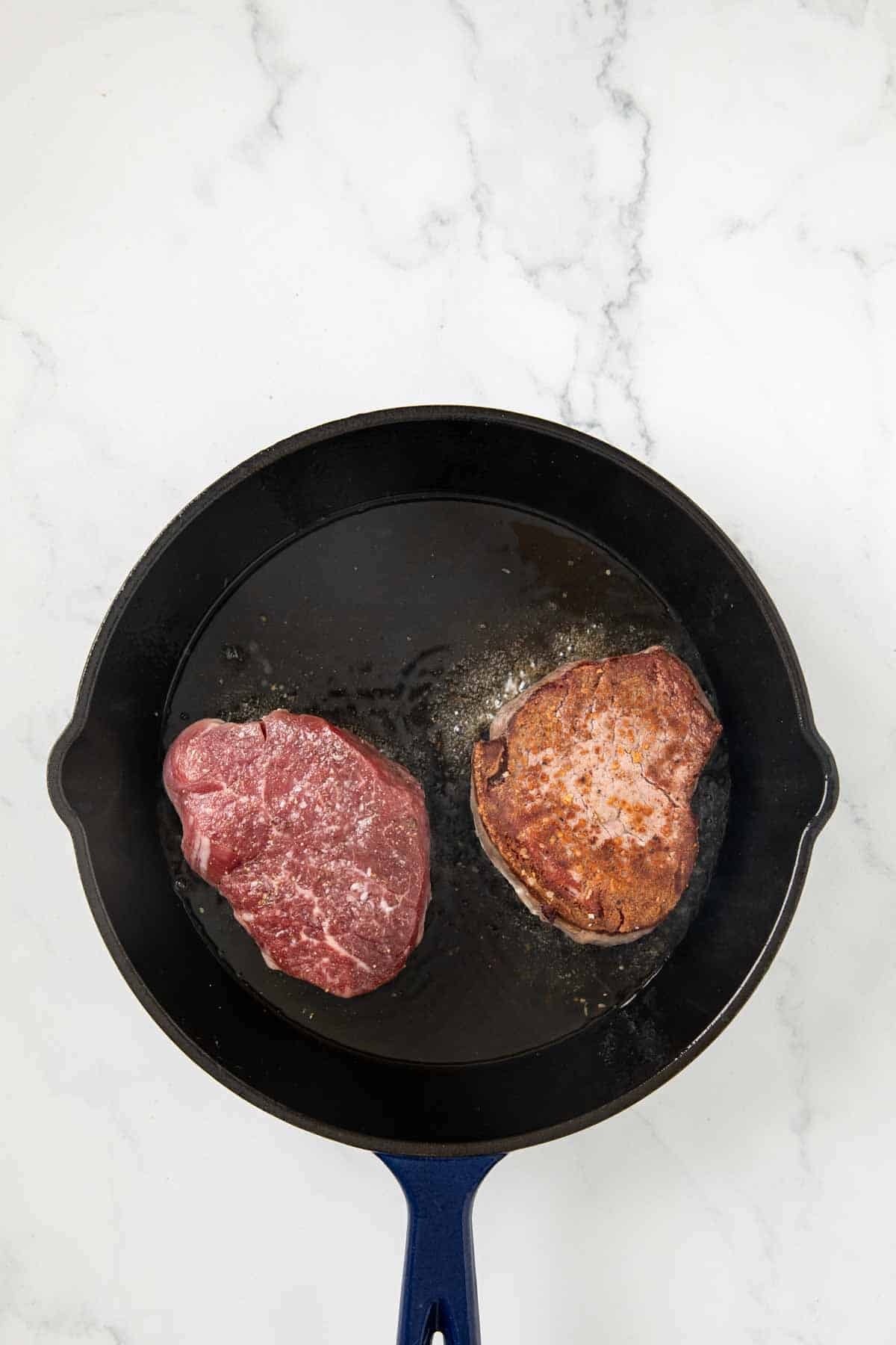 An overhead view of a cast iron skillet with two filet mignon steaks cooking - one raw and one partially cooked showing searing on one side. The pan rests on a white marble surface.