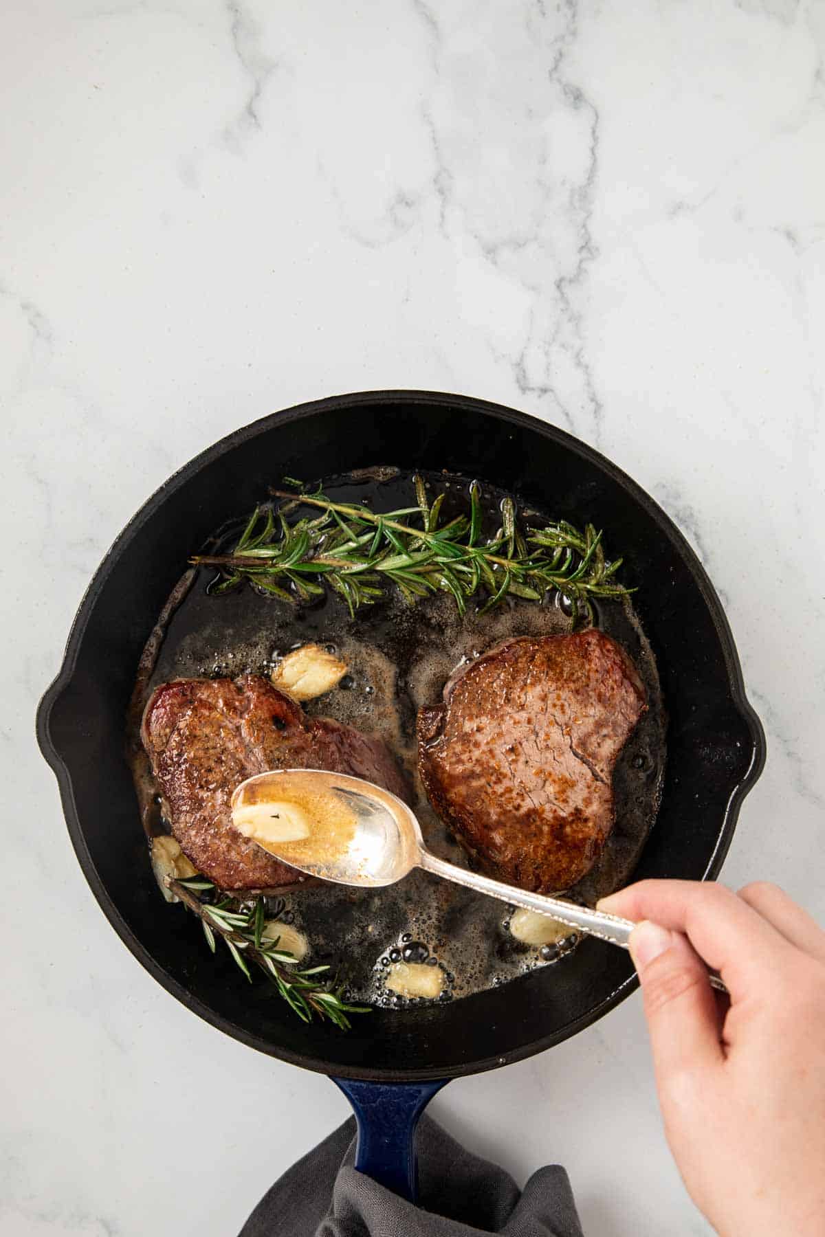 A cast iron skillet containing two seared filet mignon steaks cooking with fresh rosemary sprigs and what appears to be garlic. Someone is using a silver spoon to baste the steaks with pan juices and butter.