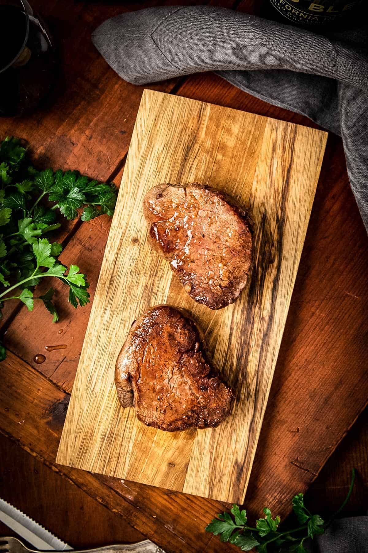 Two perfectly cooked filet mignon steaks on a wooden cutting board garnished with fresh parsley. The steaks have a caramelized exterior and sit against a rustic wooden background with a glass of red wine visible.