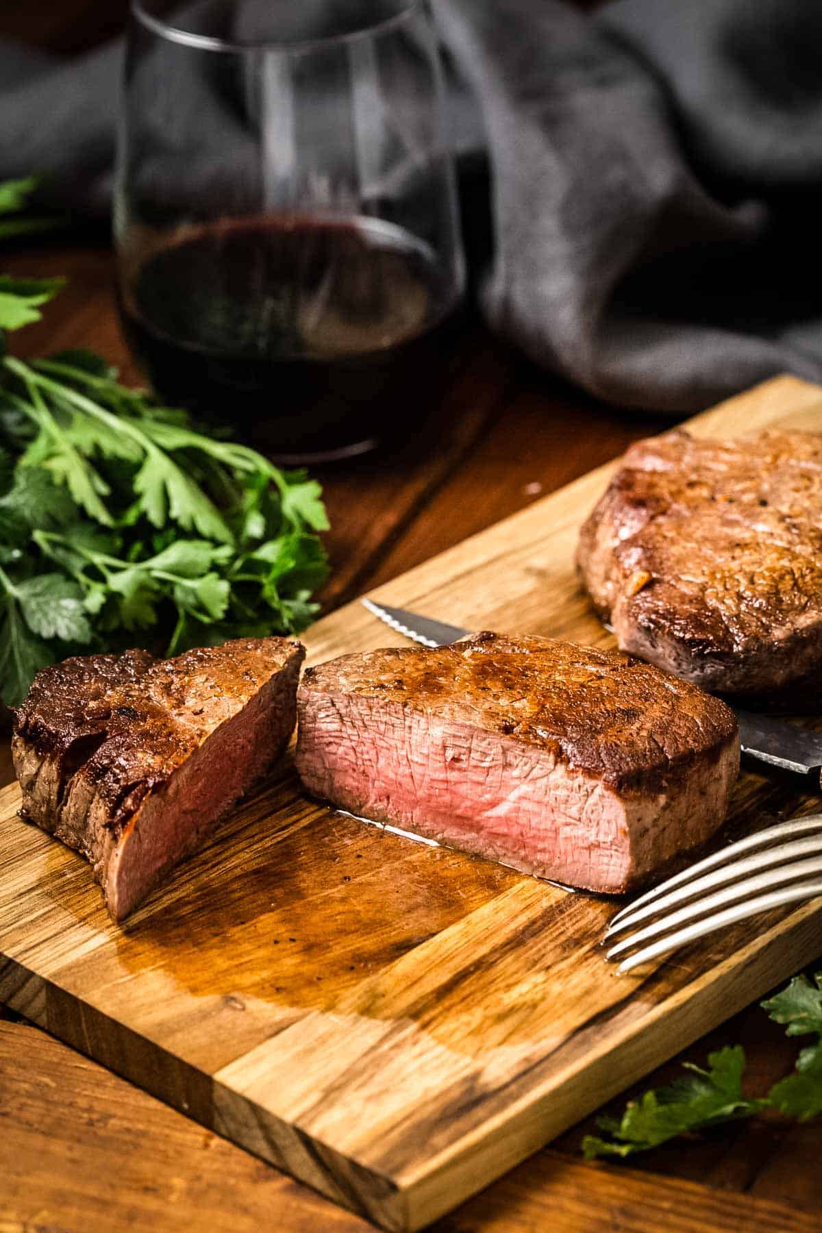 A cut-open filet mignon showing its medium-rare interior next to another whole steak on a wooden board. Fresh parsley is visible in the corner, and a serving fork rests on the board.