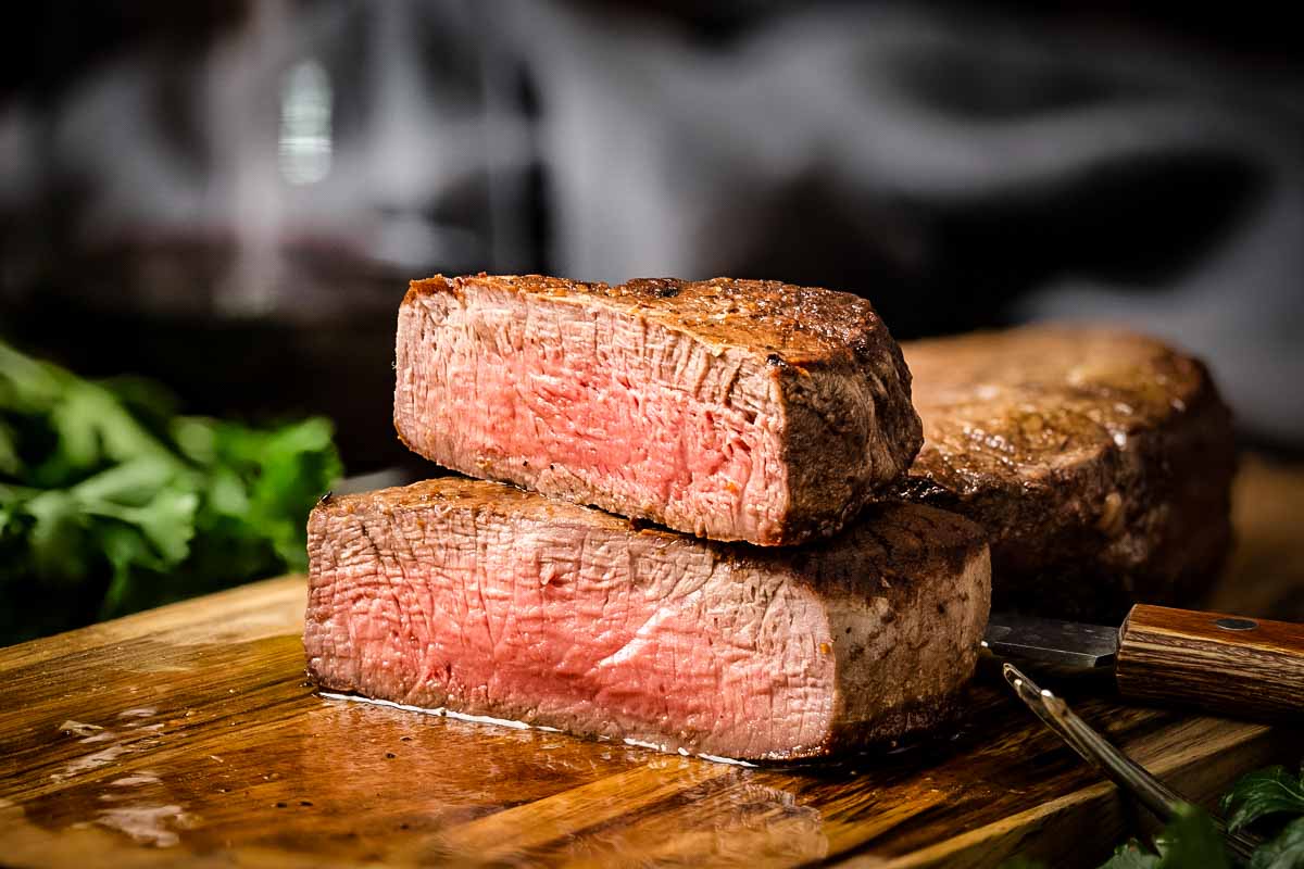 A dramatic close-up of two stacked filet mignon steaks with one cut open to display its perfectly medium-rare center. The photo captures the contrast between the seared exterior and red interior.