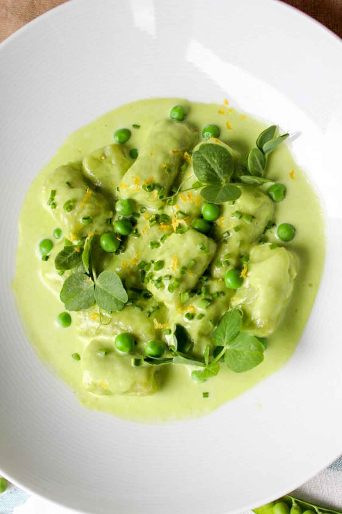 Pillowy green gnocchi served in a creamy light green sauce with fresh peas, microgreens, and orange zest on a white plate.